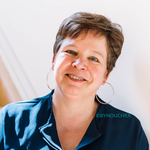 Hilde - de oprichster van Braazh: een lachende vrouw met kort kastanjebruin haar, grote oorbellen en een blauwe blouse, voor een lichte achtergrond.