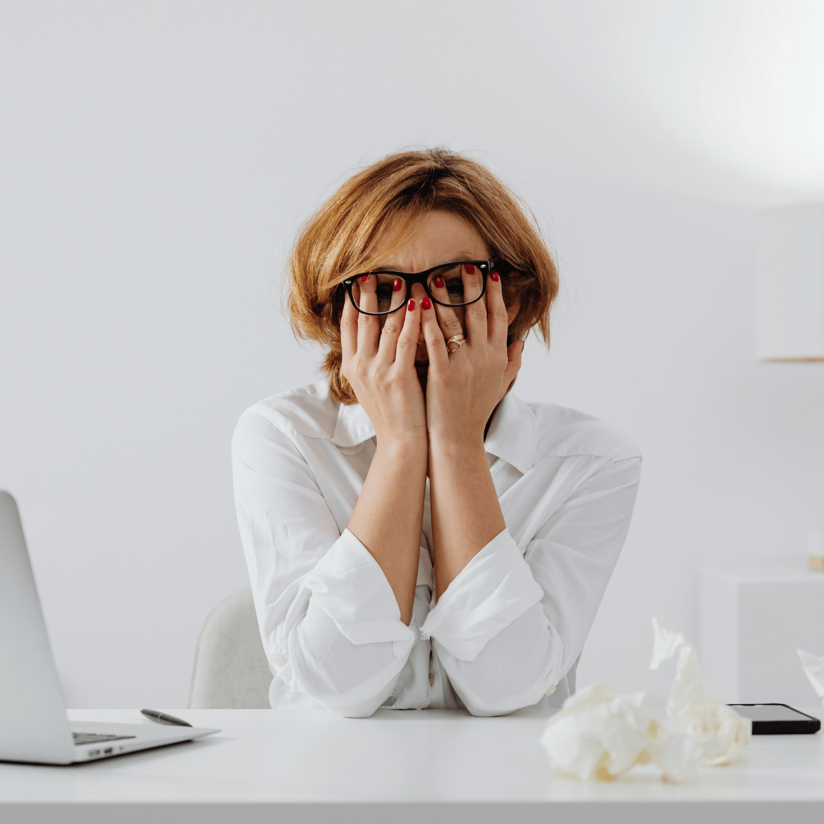 Vrouw met rood haar en zwarte glazen, zit achter een bureau, met haar handen over haar gezicht, omringd door papieren rommel. Het gaat even niet meer
