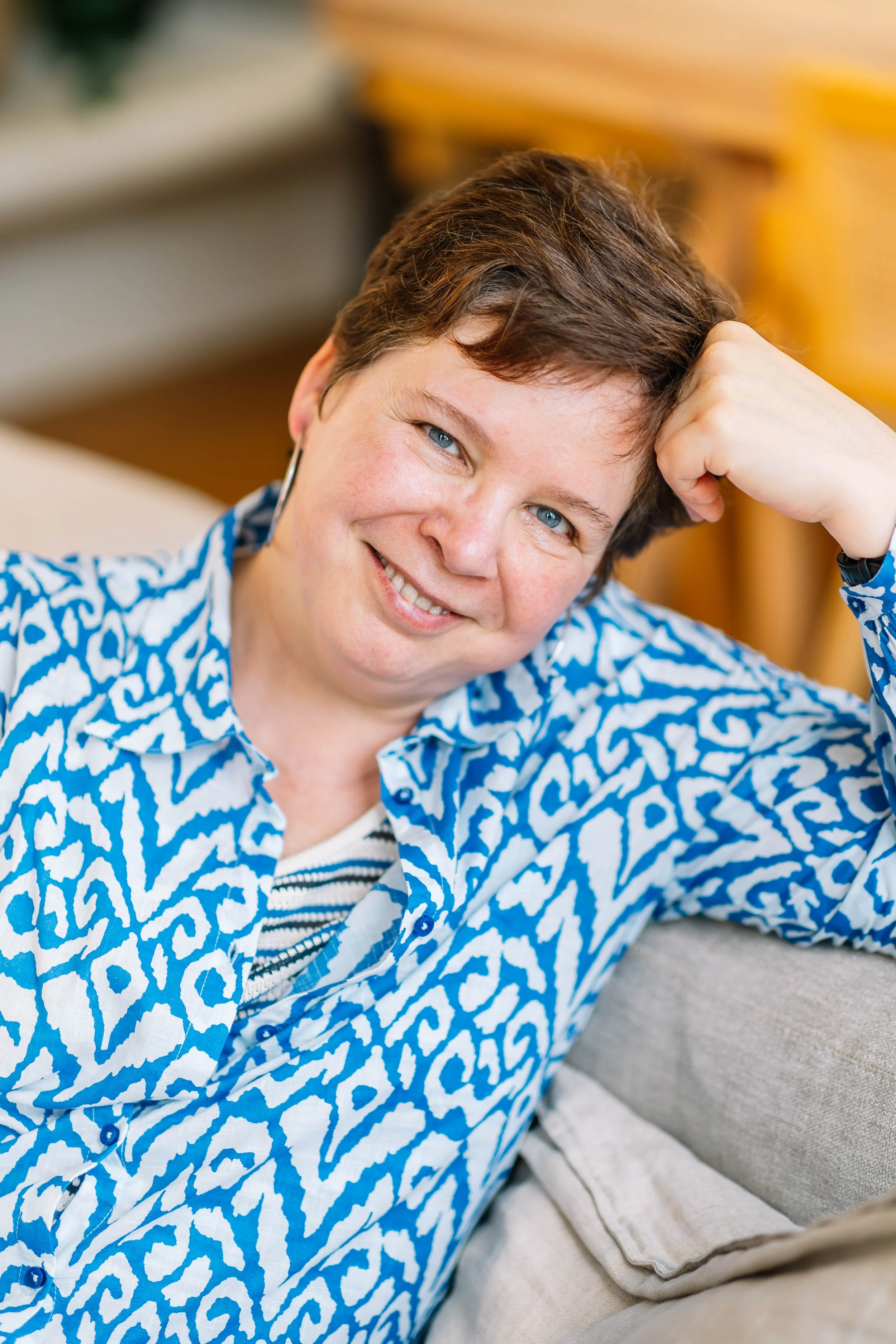 Close-up van een lachende vrouw met kort bruin haar en blauwe ogen die in een comfortabele zithoek ligt, rustend met haar onderarm op een kussen, shirt met blauwe en witte patroon.