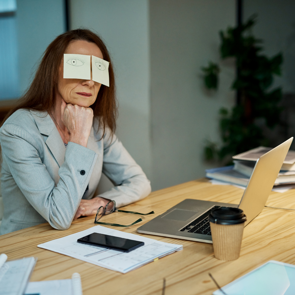 Vrouw met papieren ogen op haar gezicht zittend aan een bureau met een laptop, koffiekopje, smartphone en documenten. Ze is uitgeput maar durft het niet te tonen