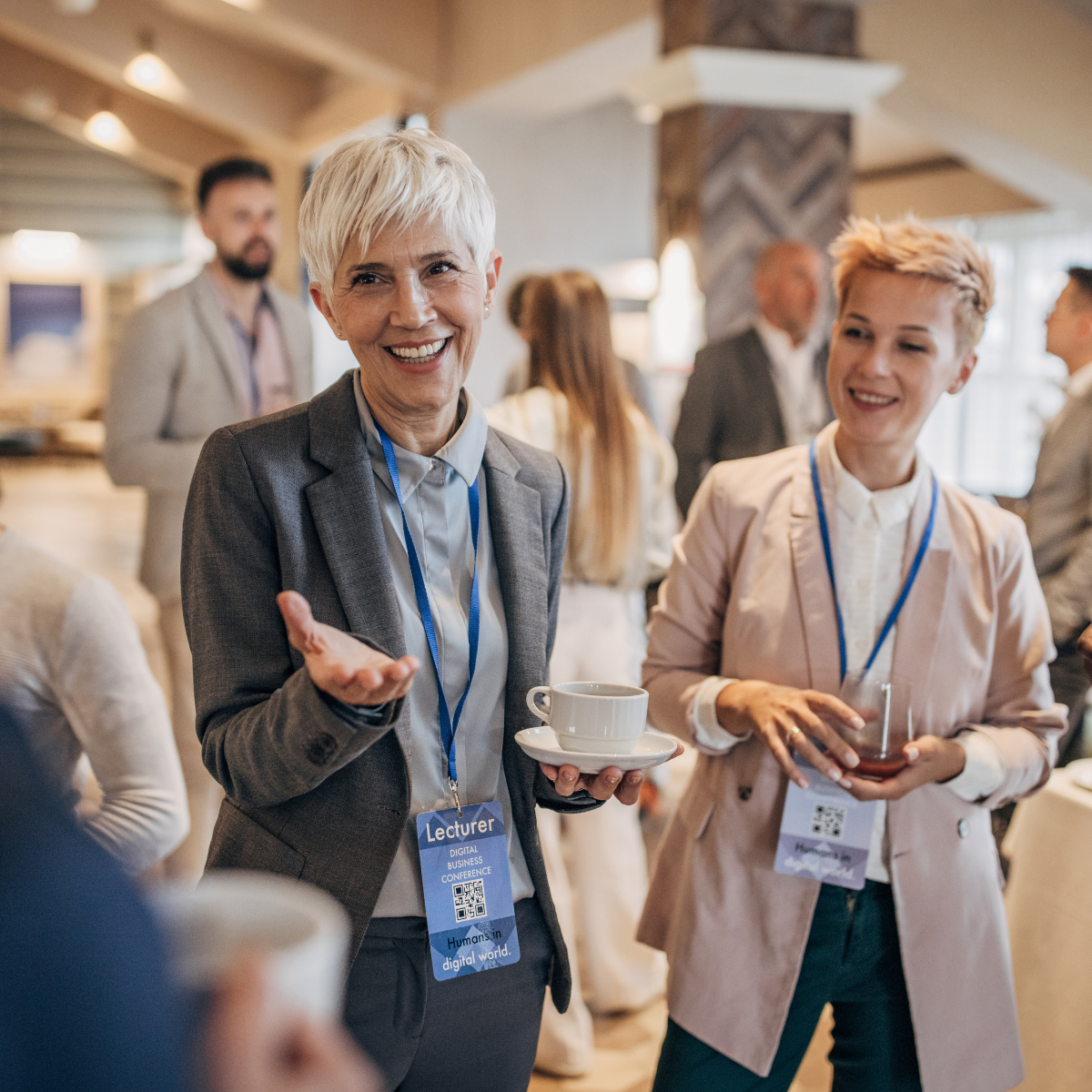 Twee vrouwen in midlife die netwerken tijdens een conferentie, één met een kopje koffie en de ander met een glas wijn, terwijl ze lachend praten.