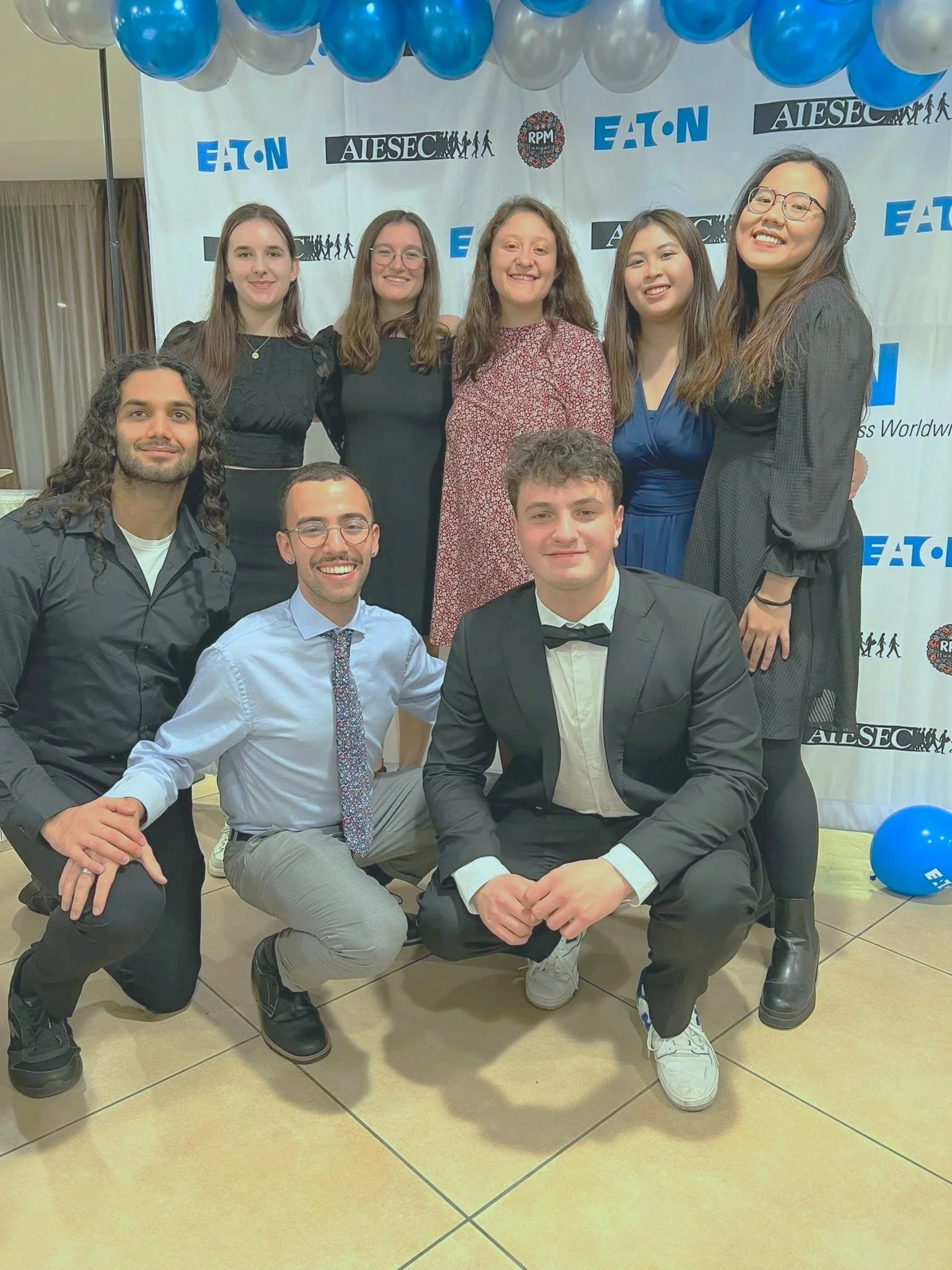 A group of ten young adults dressed in formal and semi-formal attire, posing in front of a white backdrop with event logos and balloons for a celebration or award ceremony.