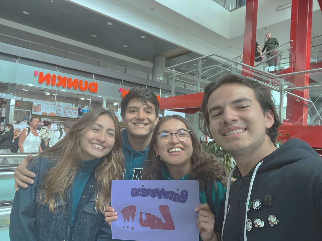 Four smiling young adults taking a selfie inside a mall, with a group holding a sign that reads 'Discover LA!!!' and a Dunkin' donut shop visible in the background.