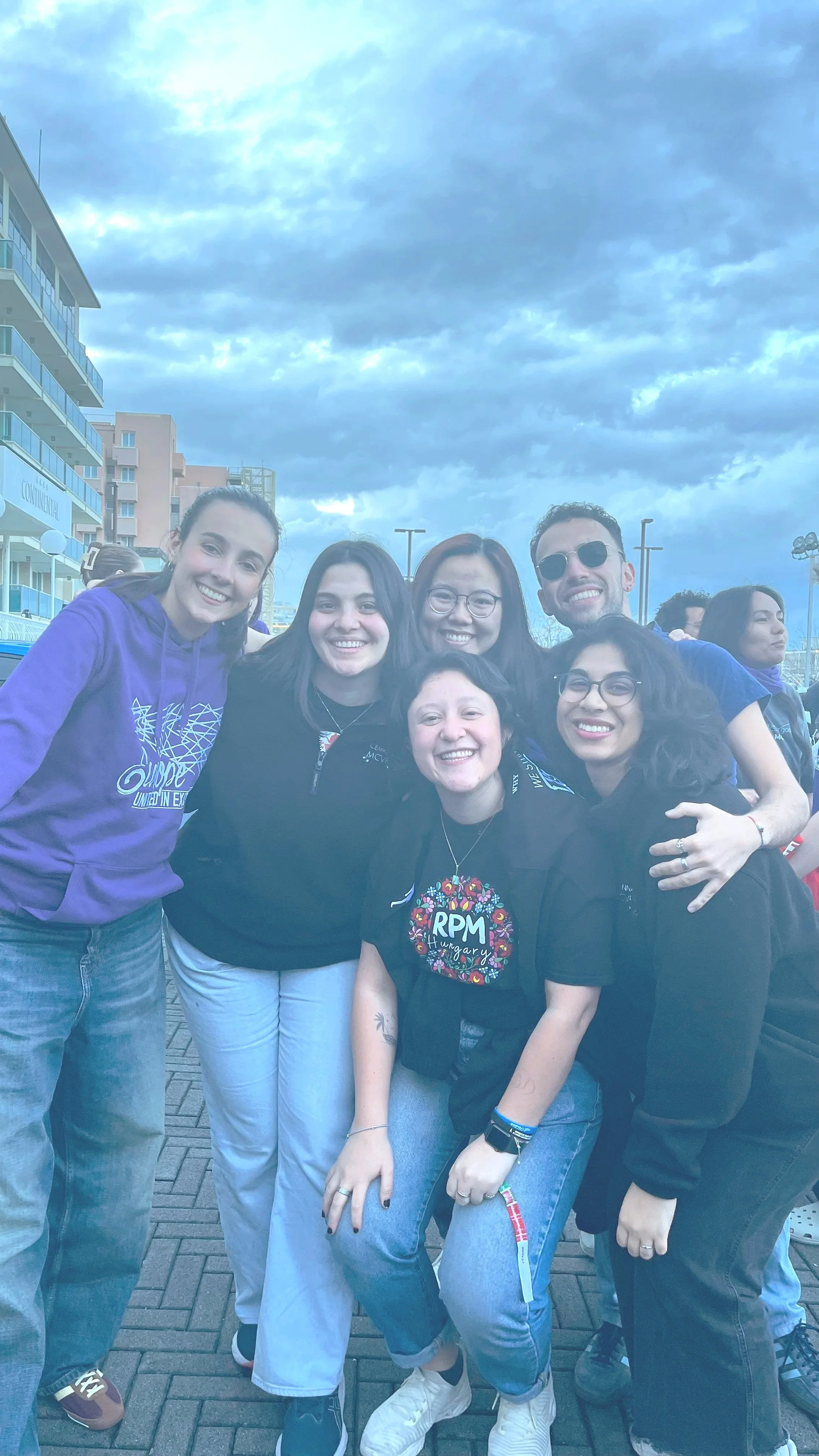 Group of smiling people standing outdoors, hugging, with a cloudy sky and buildings in the background.