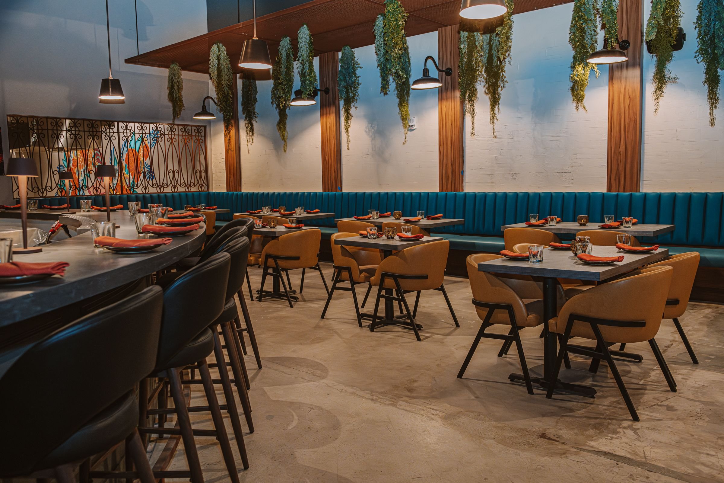 Contemporary restaurant interior with modern furniture, teal banquette seating, and hanging plants. Tables set with red napkins and glasses.
