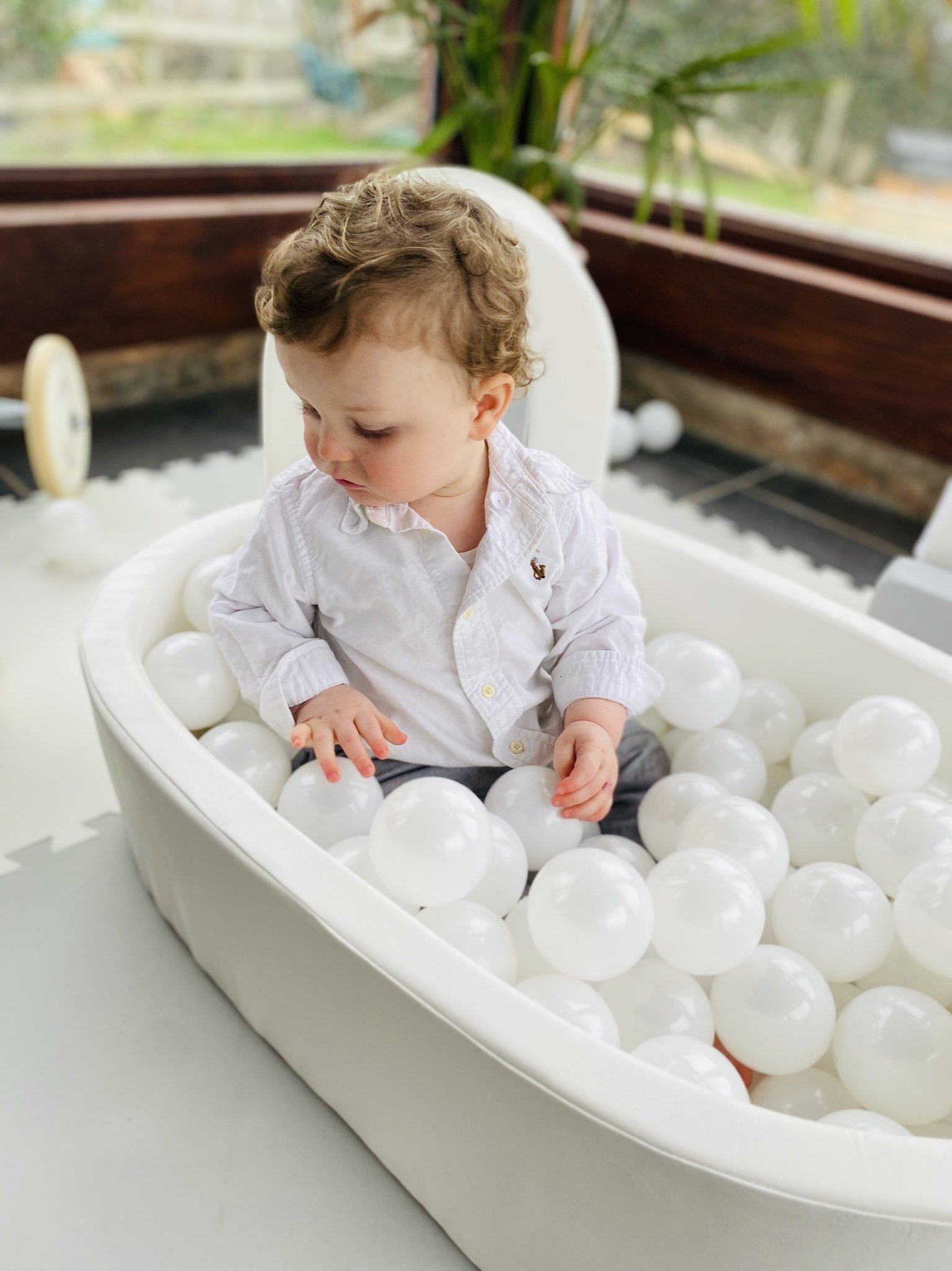 Young child with curly hair sitting in a white ball pit filled with white plastic balls.