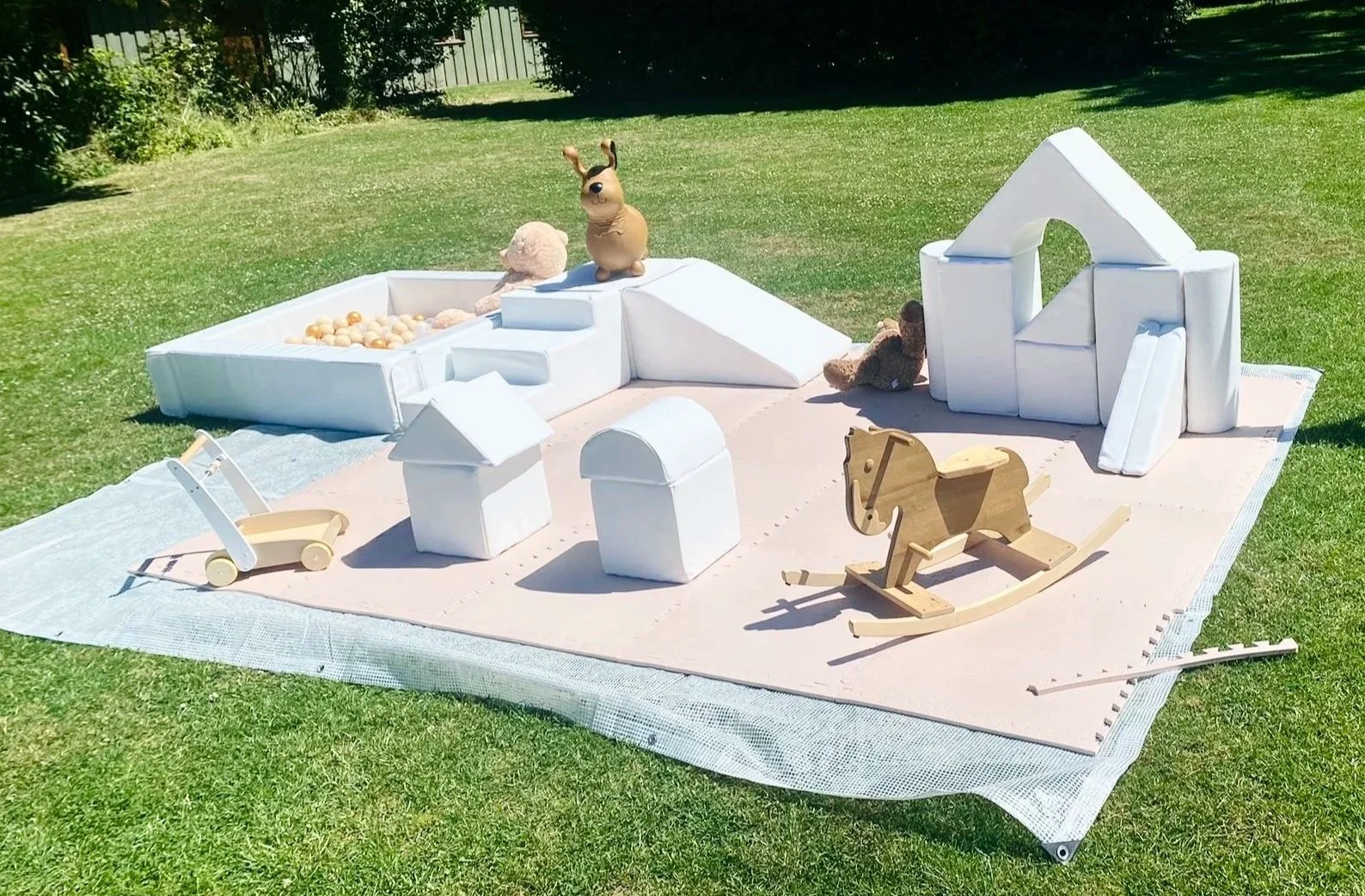 A small outdoor baby play area on grass, with soft white structures resembling a house and a slide, wooden toys including a rocking horse and a stroller, and plush toys.