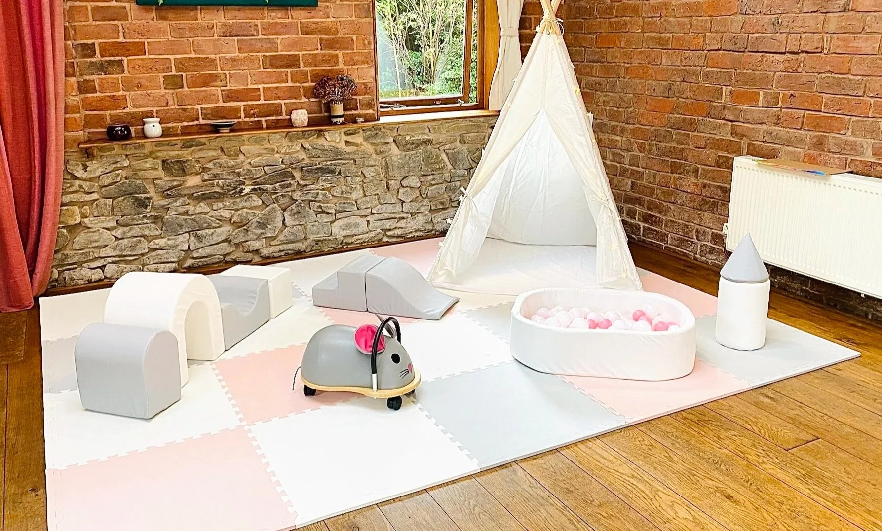 Children's play area with a small white tent, ball pit, toy mouse suitcase, foam climbing blocks, and foam tunnels on a pink and white play mat, set against a brick wall with a window.