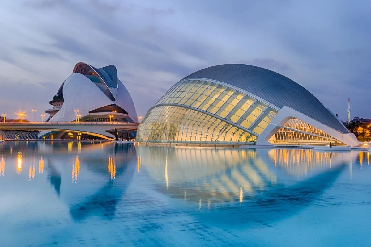 Architectural photograph of the City of Arts and Sciences in Valencia, Spain, featuring modern white structures, a reflective water body, and a cloudy sky.