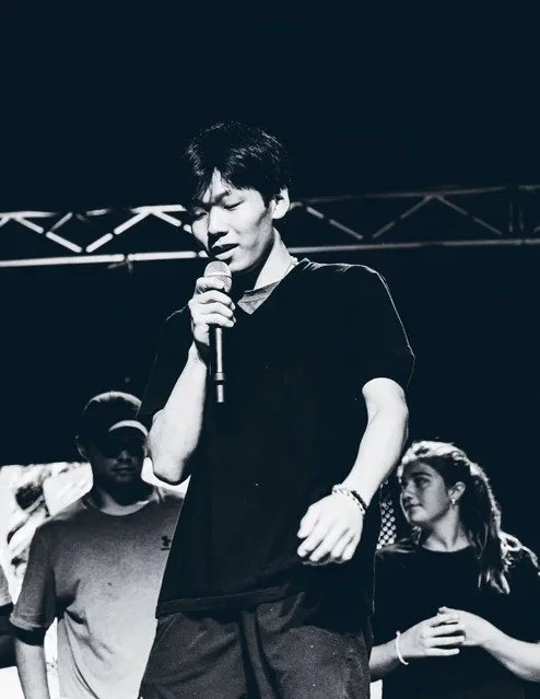 Young man singing into a microphone on stage with two other people in the background, one wearing glasses and a cap, under stage lights.