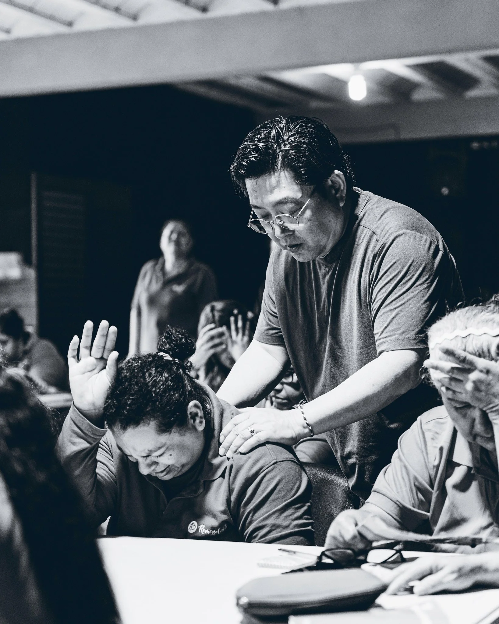 Black and white photo of a man with glasses leaning over a table, engaging with others. Several people in the background, some with hands raised, in what appears to be a group activity or discussion.