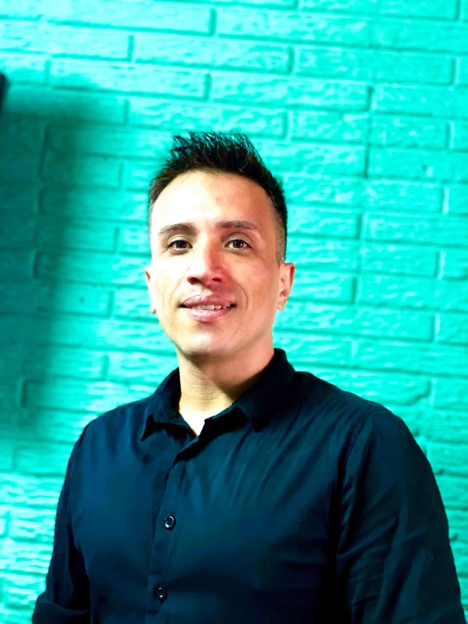 A young man with short dark hair, wearing a black collared shirt, standing in front of a turquoise brick wall.