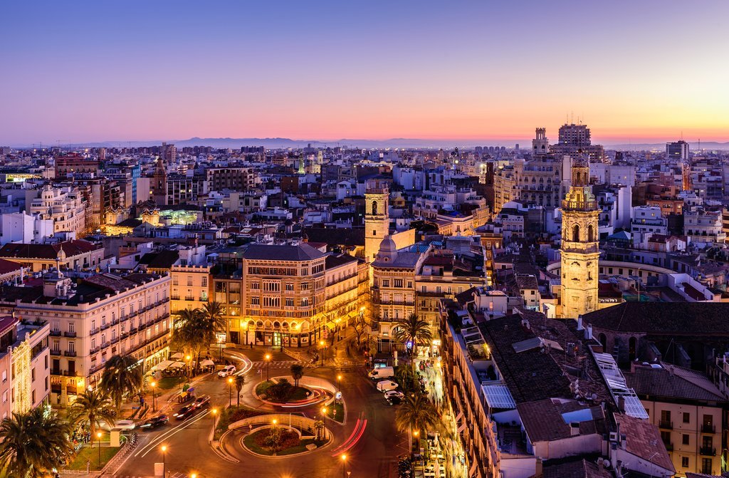 Cityscape at sunset with historic buildings, palm trees, and a roundabout in the foreground.