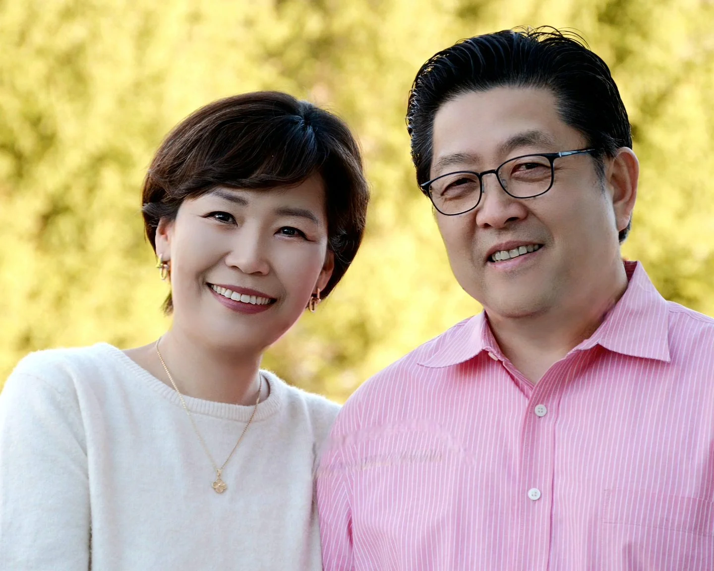 A smiling woman and a man standing close outdoors against a blurred background of trees, both dressed casually.