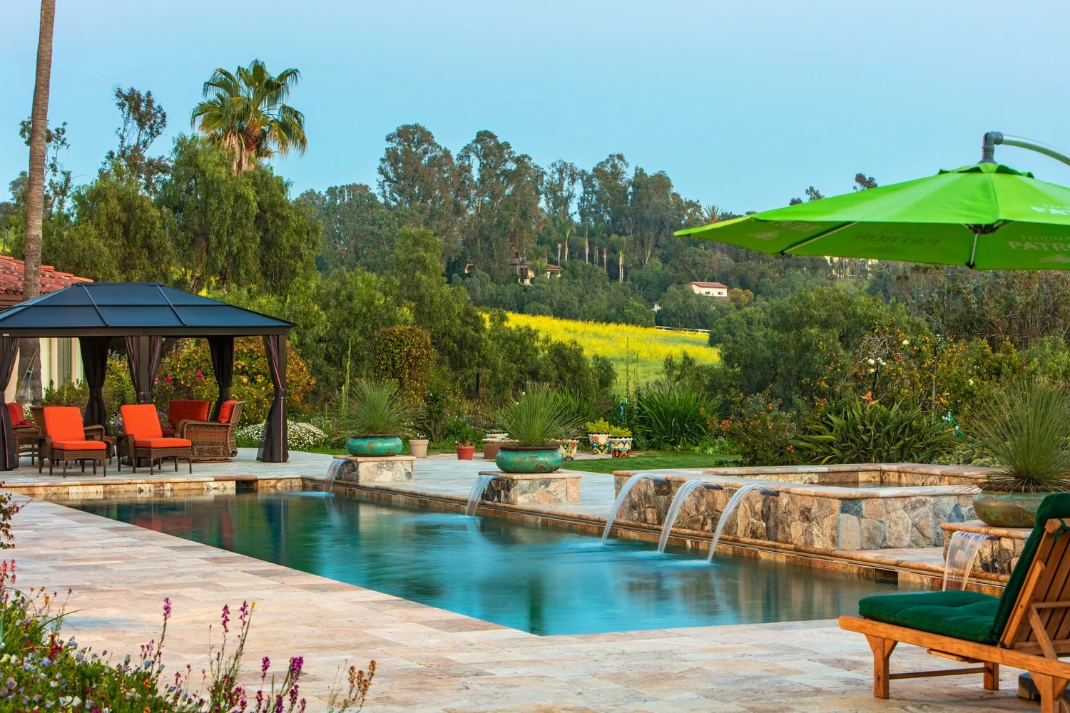 Luxurious backyard with pool, water features, gazebo with red-orange chairs, green umbrella, and lush landscaping.