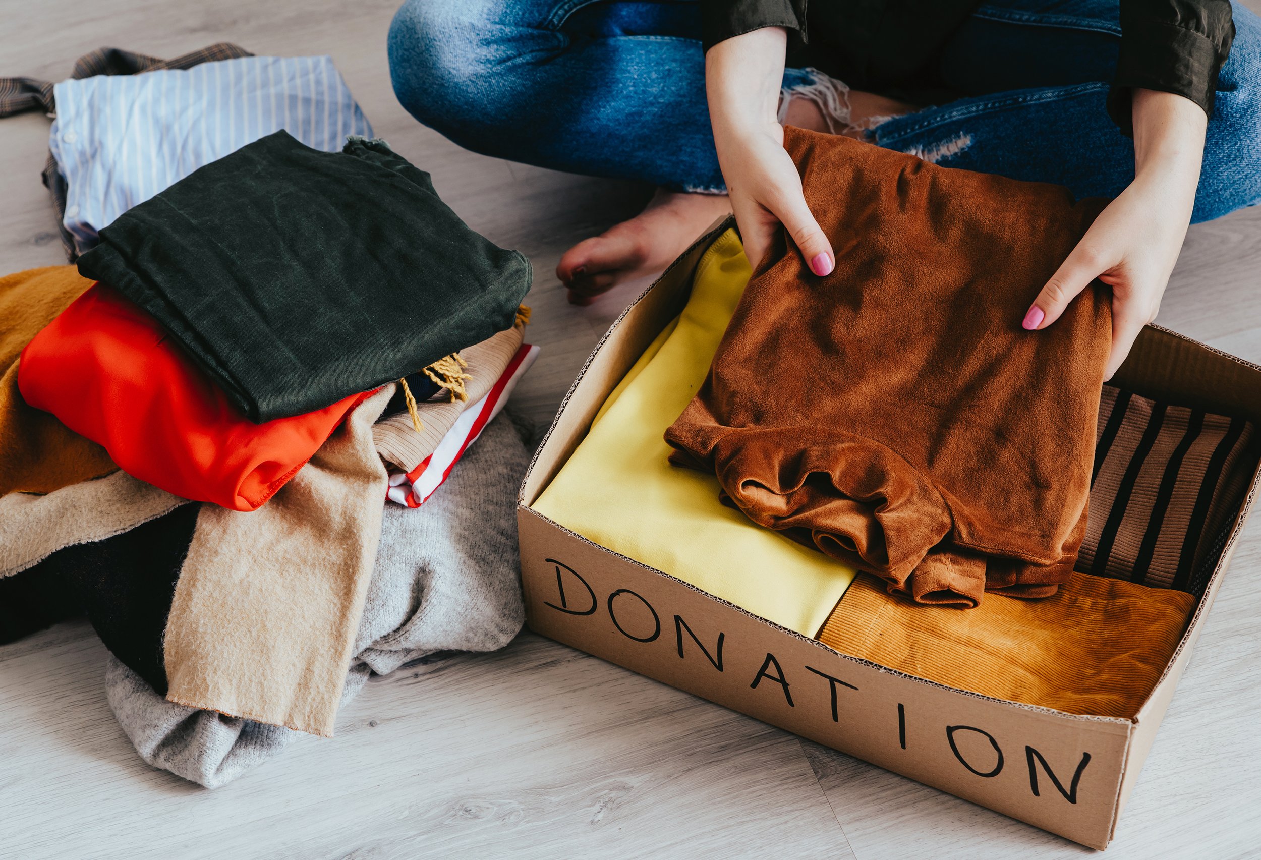 Person packing clothes into a donation box on the floor, with folded clothing nearby.