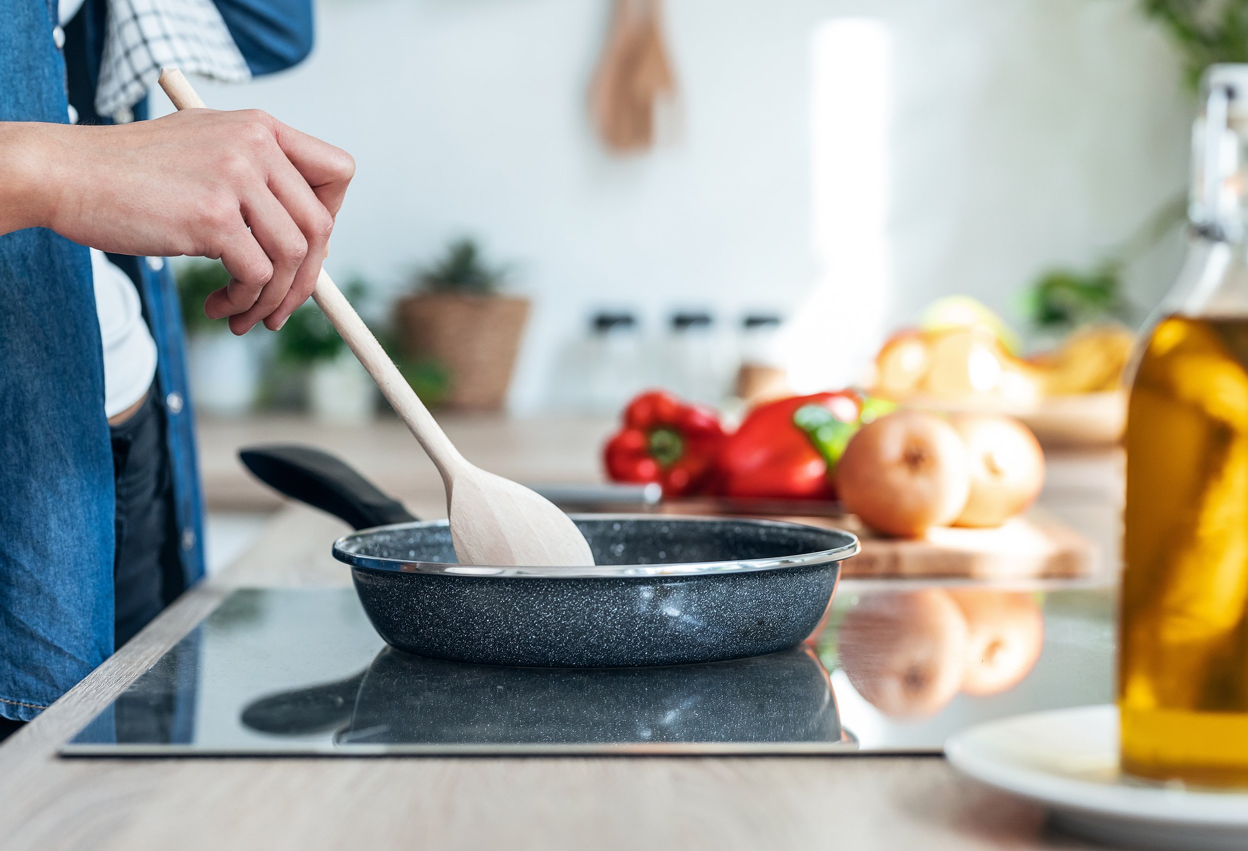 Person stirring a pan on a stovetop with a wooden spoon, surrounded by vegetables and a bottle of olive oil.