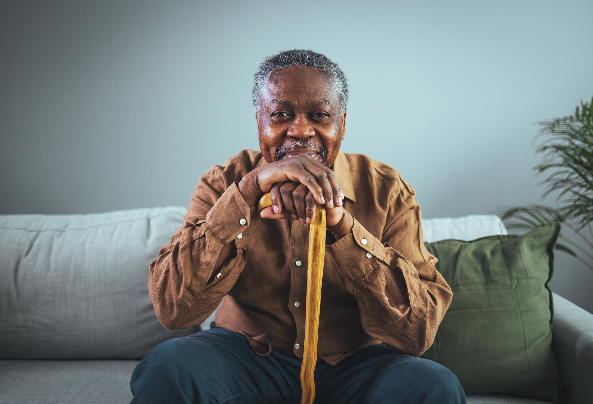 Elderly man sitting on couch with a cane, smiling and wearing a brown shirt.
