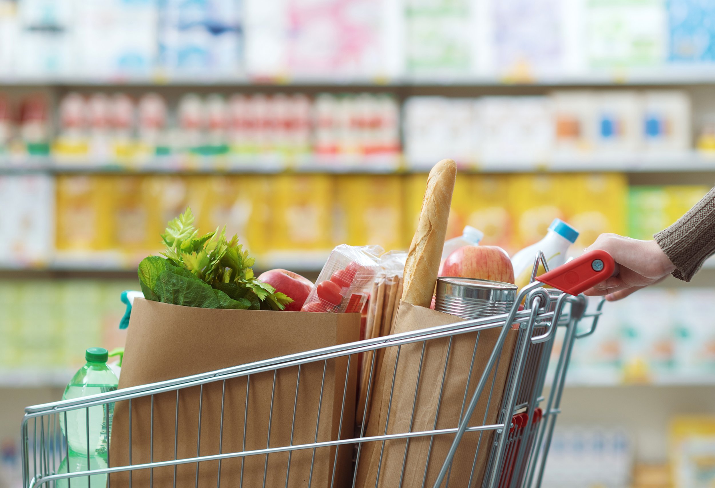 Shopping cart filled with groceries like a baguette, lettuce, apples, and canned goods in a supermarket aisle.