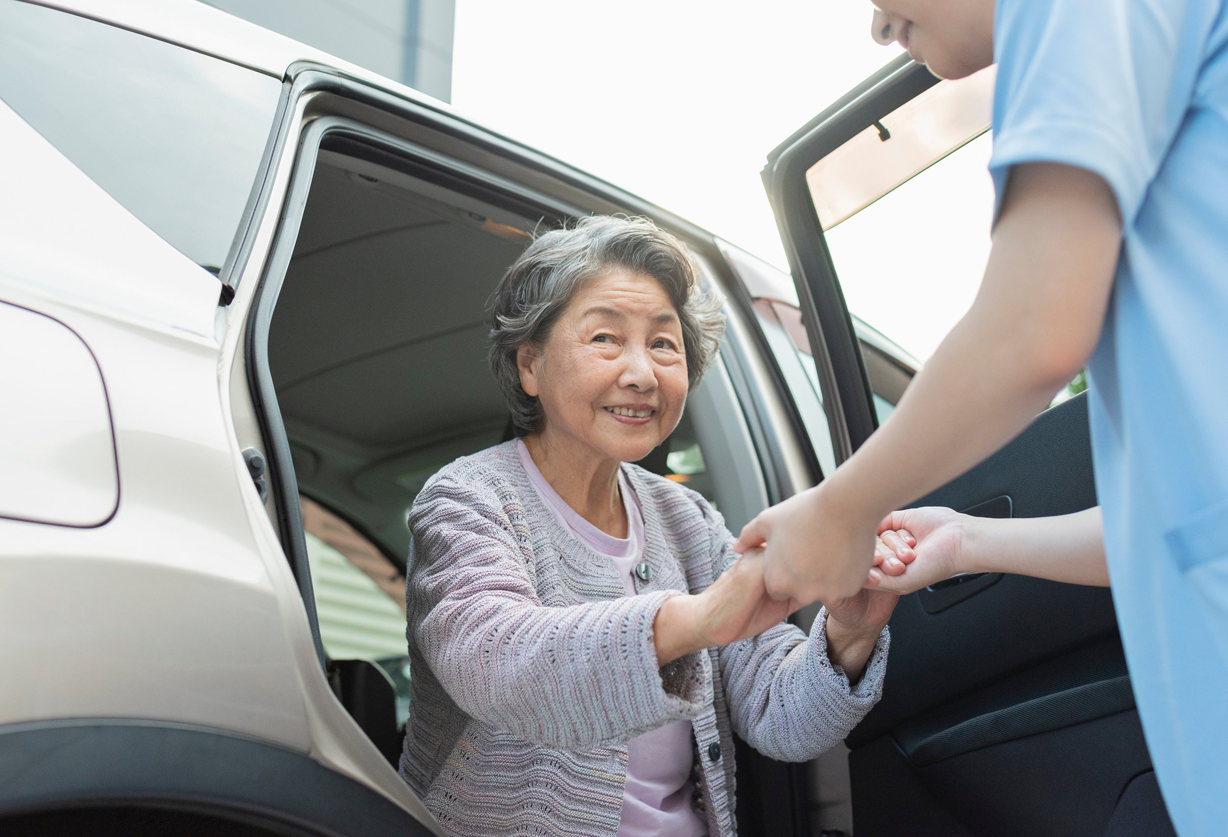 Elderly woman being assisted out of a car by a caregiver in a blue uniform.