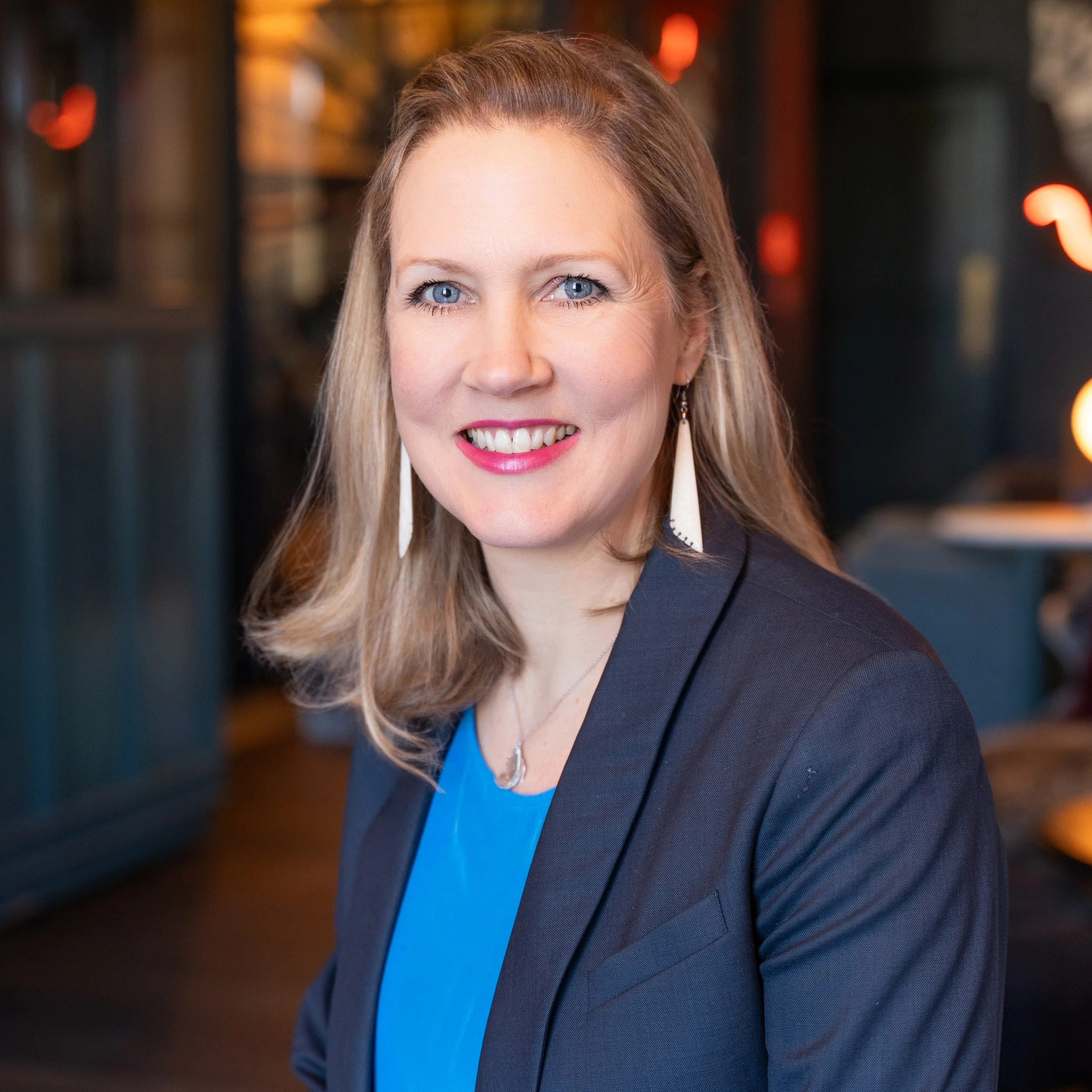 Woman smiling in a professional setting wearing a blue top and blazer, with long earrings and medium-length blonde hair.