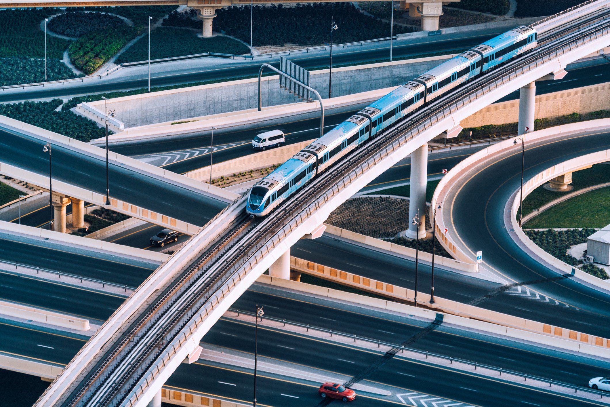 Aerial view of a modern elevated train on curving tracks over a multi-level highway interchange with cars below.