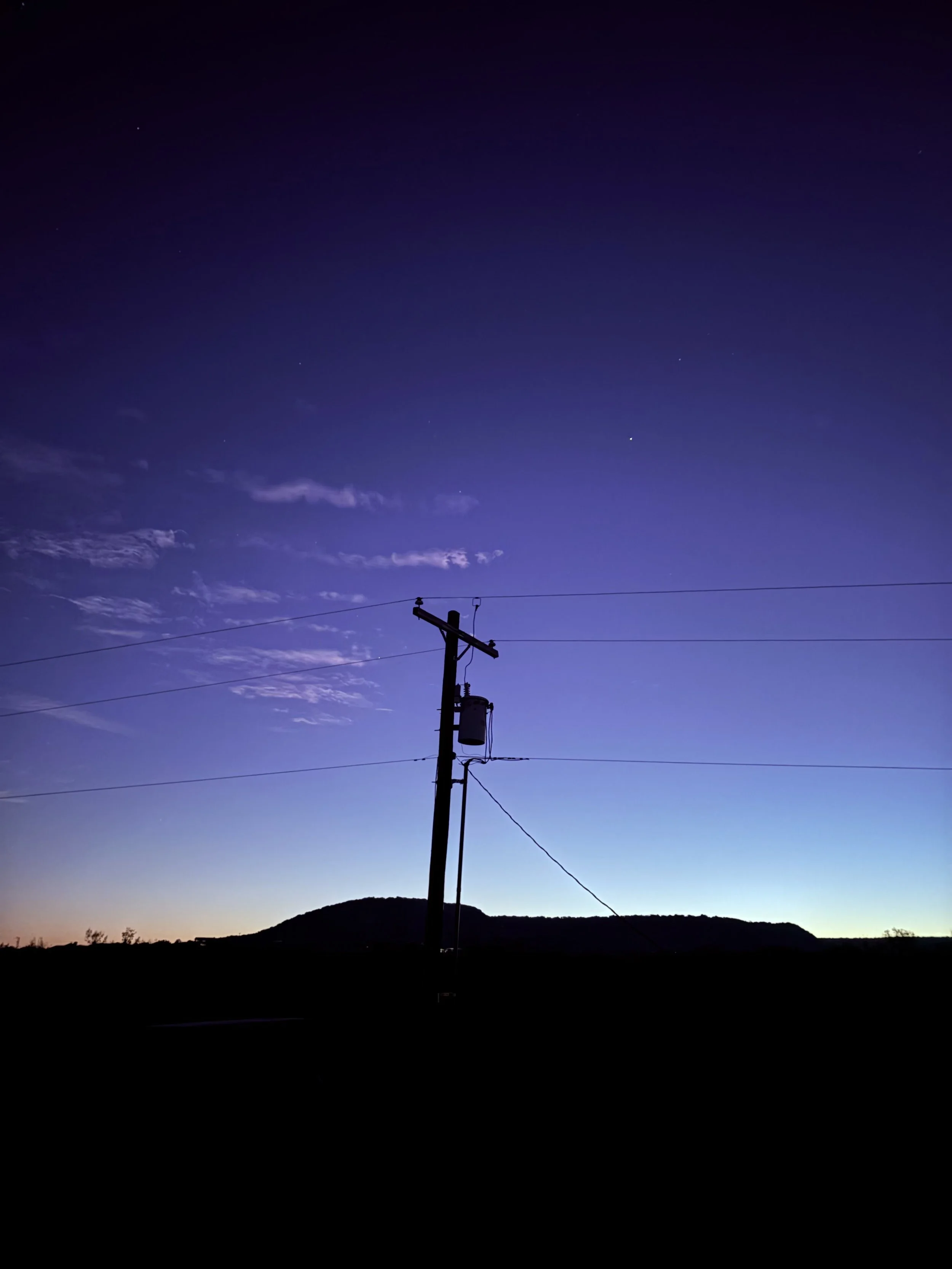 Silhouette of a utility pole with power lines against a twilight sky with a mountain in the background.