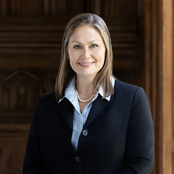 Professional woman with shoulder-length brown hair wearing a dark blazer, light blue shirt, and pearl necklace, standing indoors against a wooden background.
