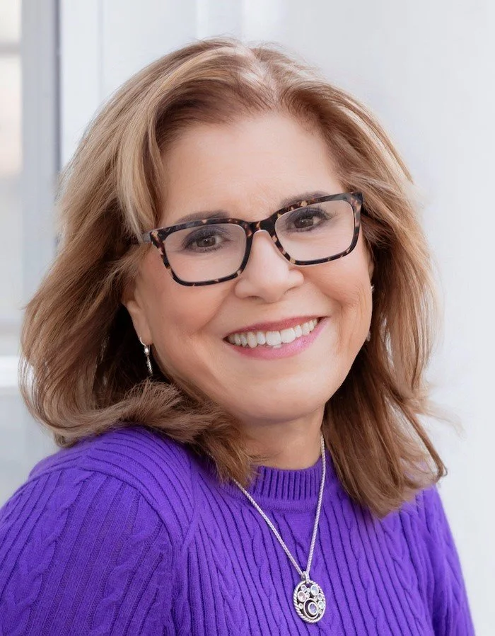 A smiling woman with shoulder-length reddish-brown hair, wearing glasses with tortoiseshell frames, a purple knit sweater, and a silver necklace, standing indoors near a window.