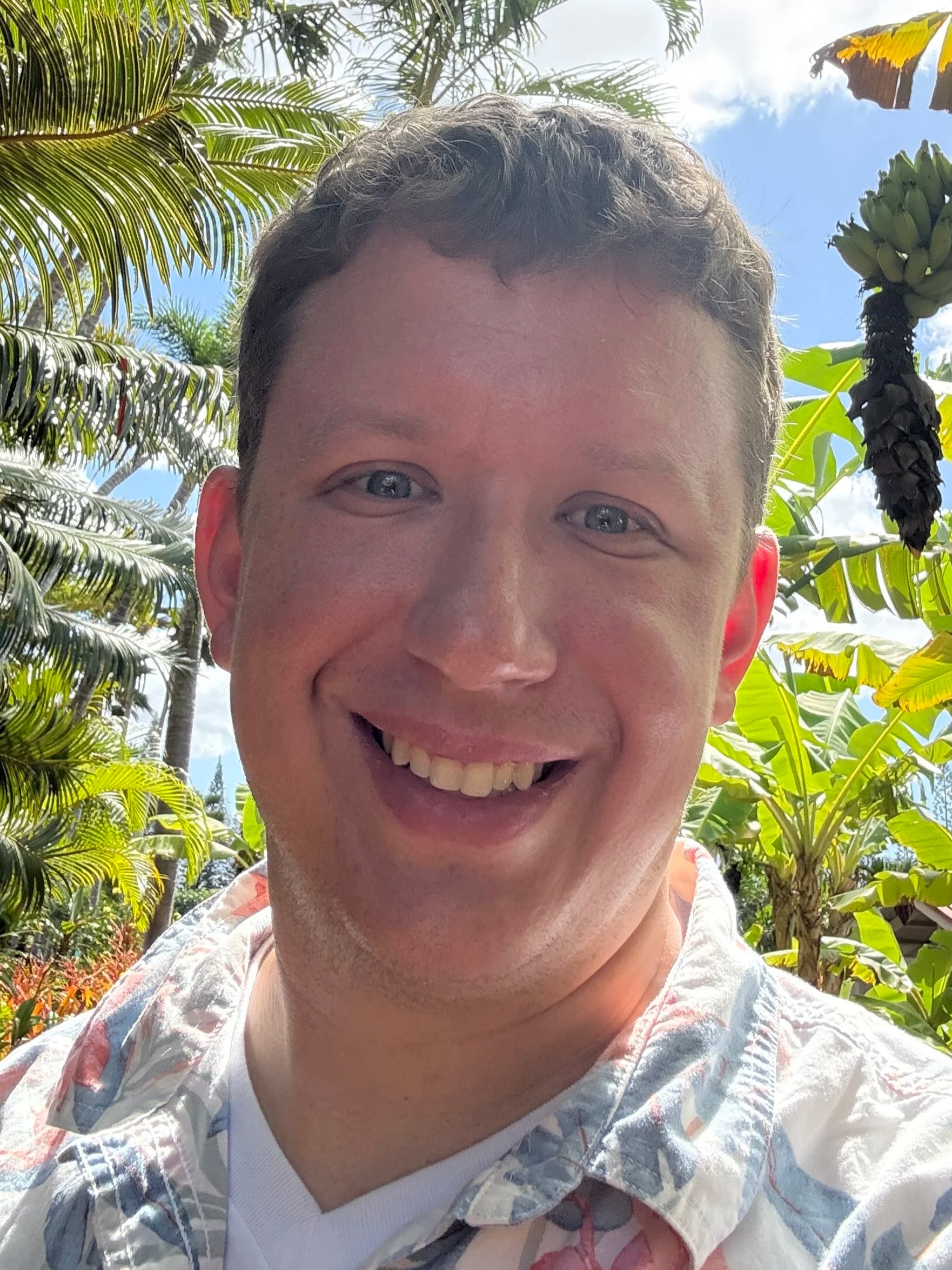Smiling man with short hair, wearing a white floral shirt, standing outdoors with tropical plants and a blue sky in the background.