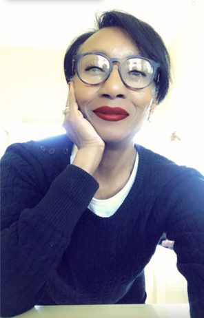Woman with short dark hair wearing glasses and a navy blue sweater, posing with her hand on her cheek and smiling at the camera.