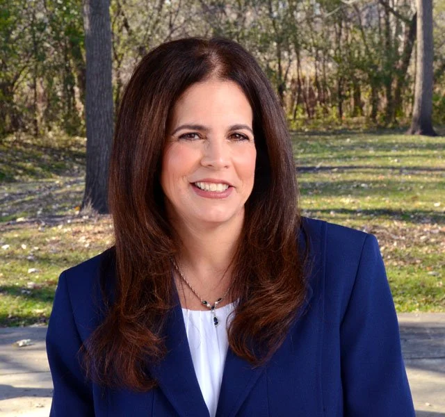 A woman with long brown hair smiling outdoors in a park setting with trees and grass, wearing a navy blue blazer over a white top.