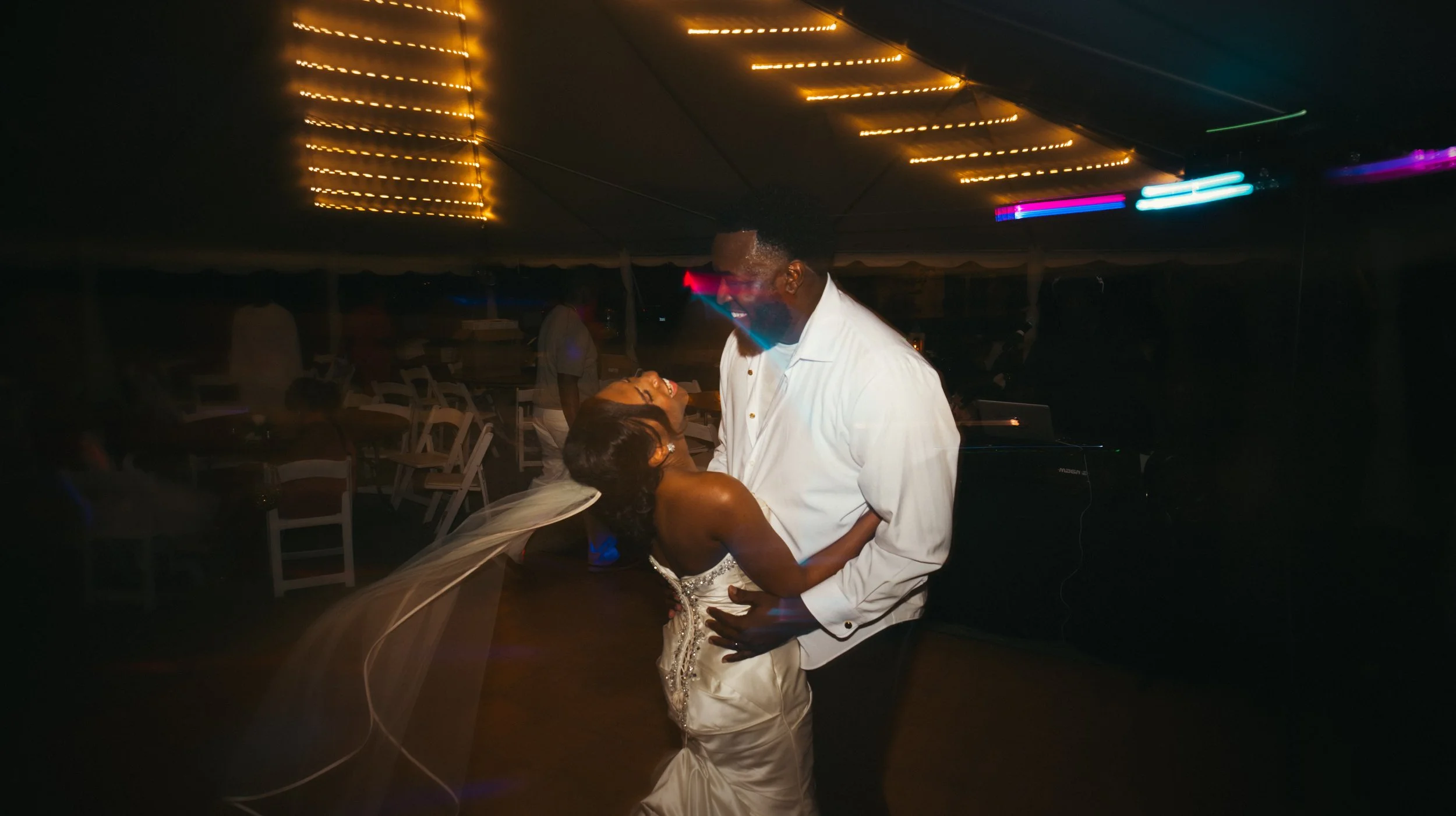 A couple dancing closely at a nighttime celebration inside a tent with string lights. The woman is wearing a wedding dress and veil, and the man is dressed in a white shirt, smiling and looking at each other.