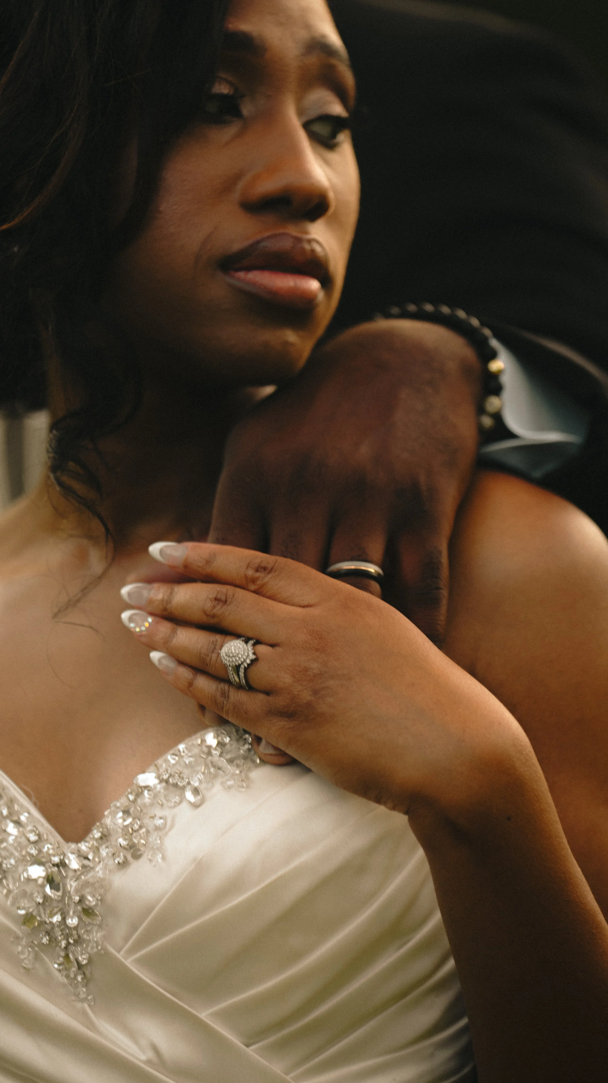 A close-up of a woman in a white dress with rhinestone embellishments, gently holding her hand on her chest, with a man's hand resting on her shoulder in a loving embrace, both showing rings.