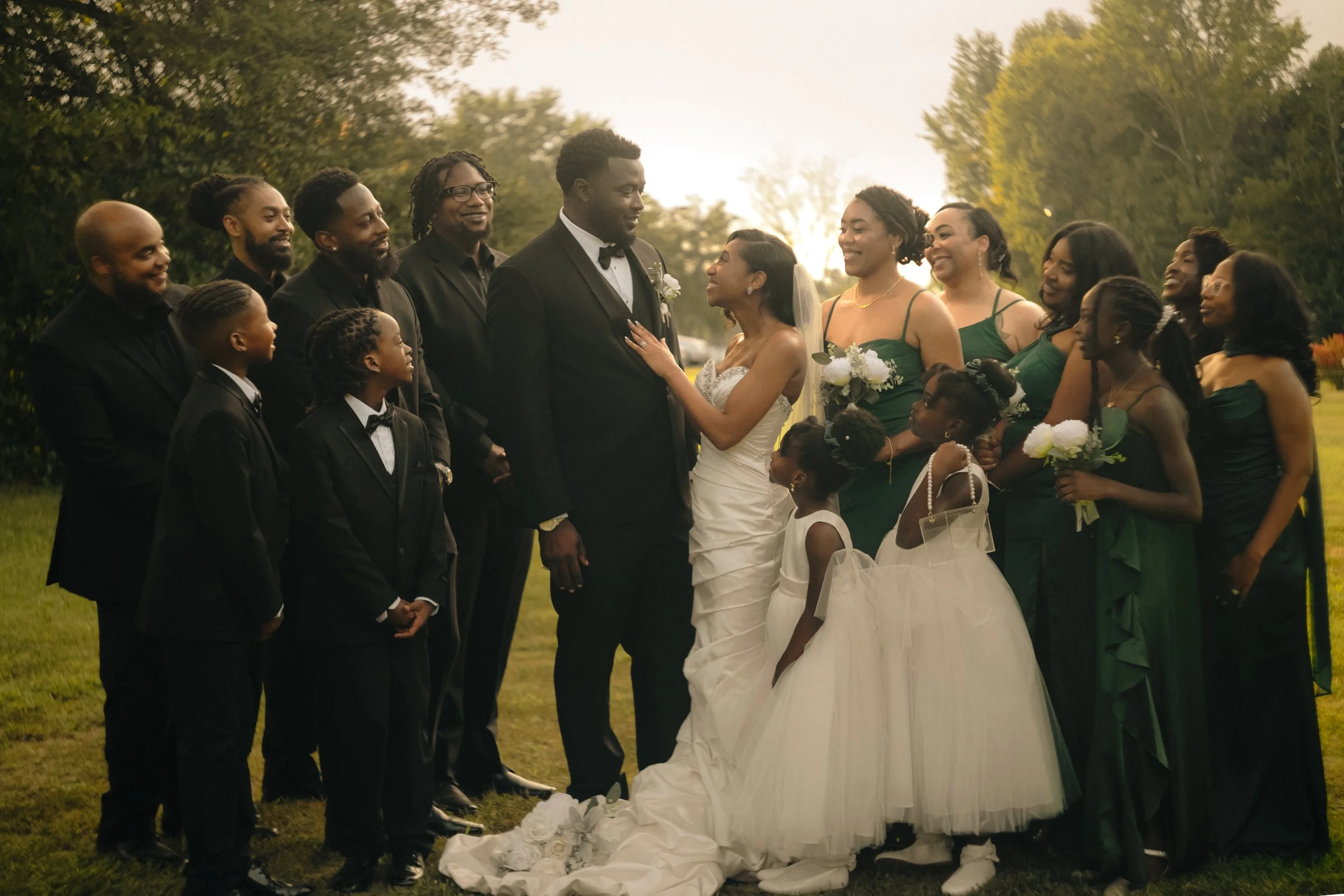 A wedding party outdoors with the bride and groom in the center, surrounded by family and friends, during sunset.
