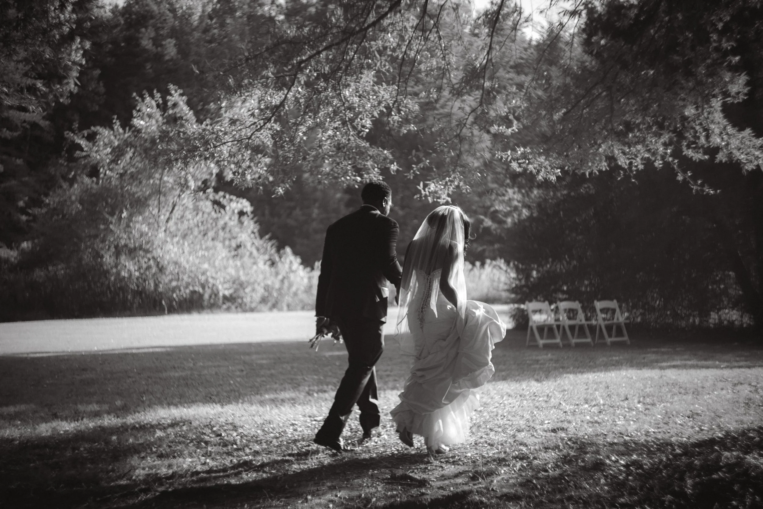 A black and white photo of a bride and groom walking together outdoors, the bride holding up her wedding gown and the groom holding a bouquet, surrounded by trees and chairs in the background.