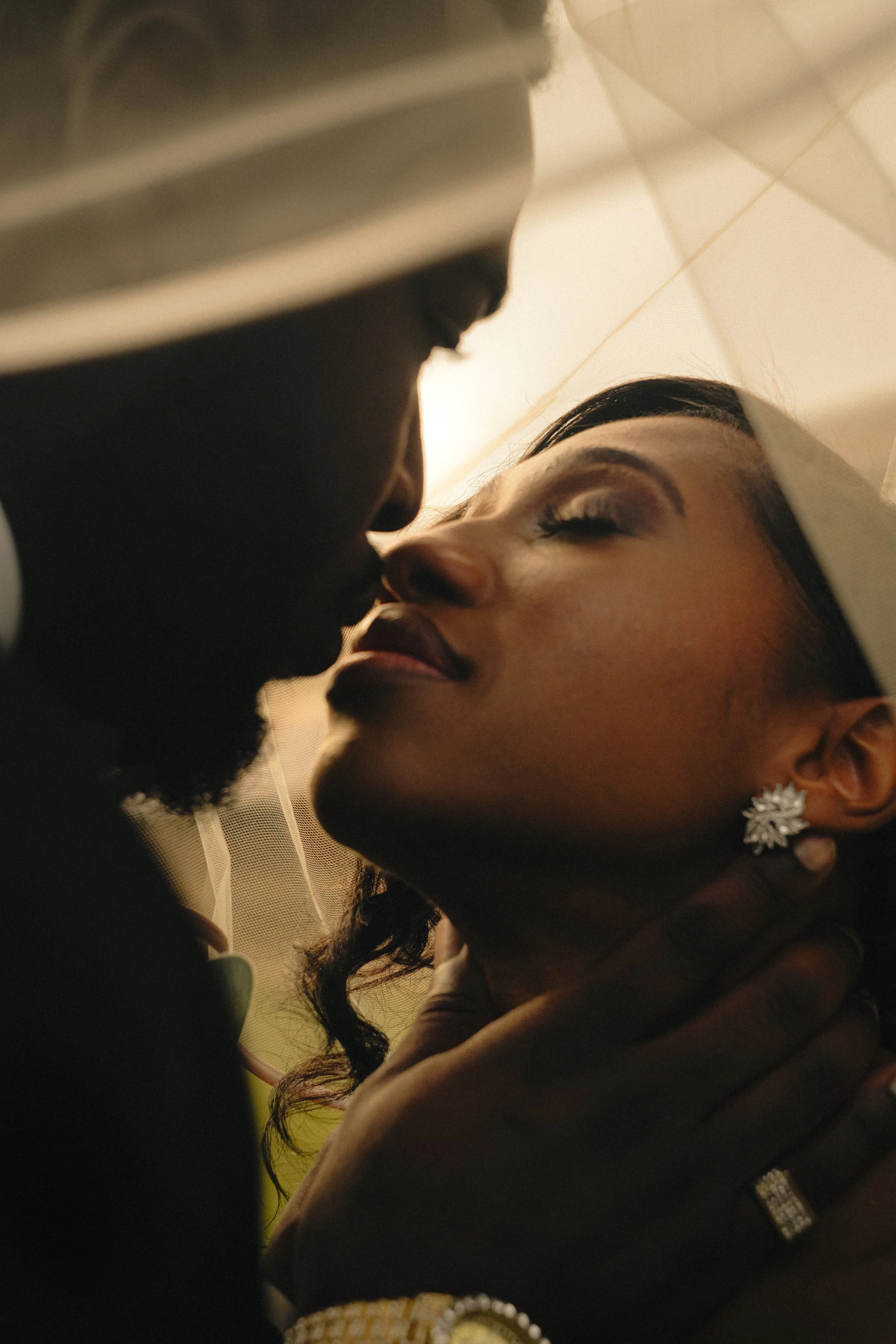 A close-up of a couple about to kiss, with the man's hand gently holding the woman's neck. The woman is wearing elegant earrings and a ring, and her eyes are closed in an intimate moment.