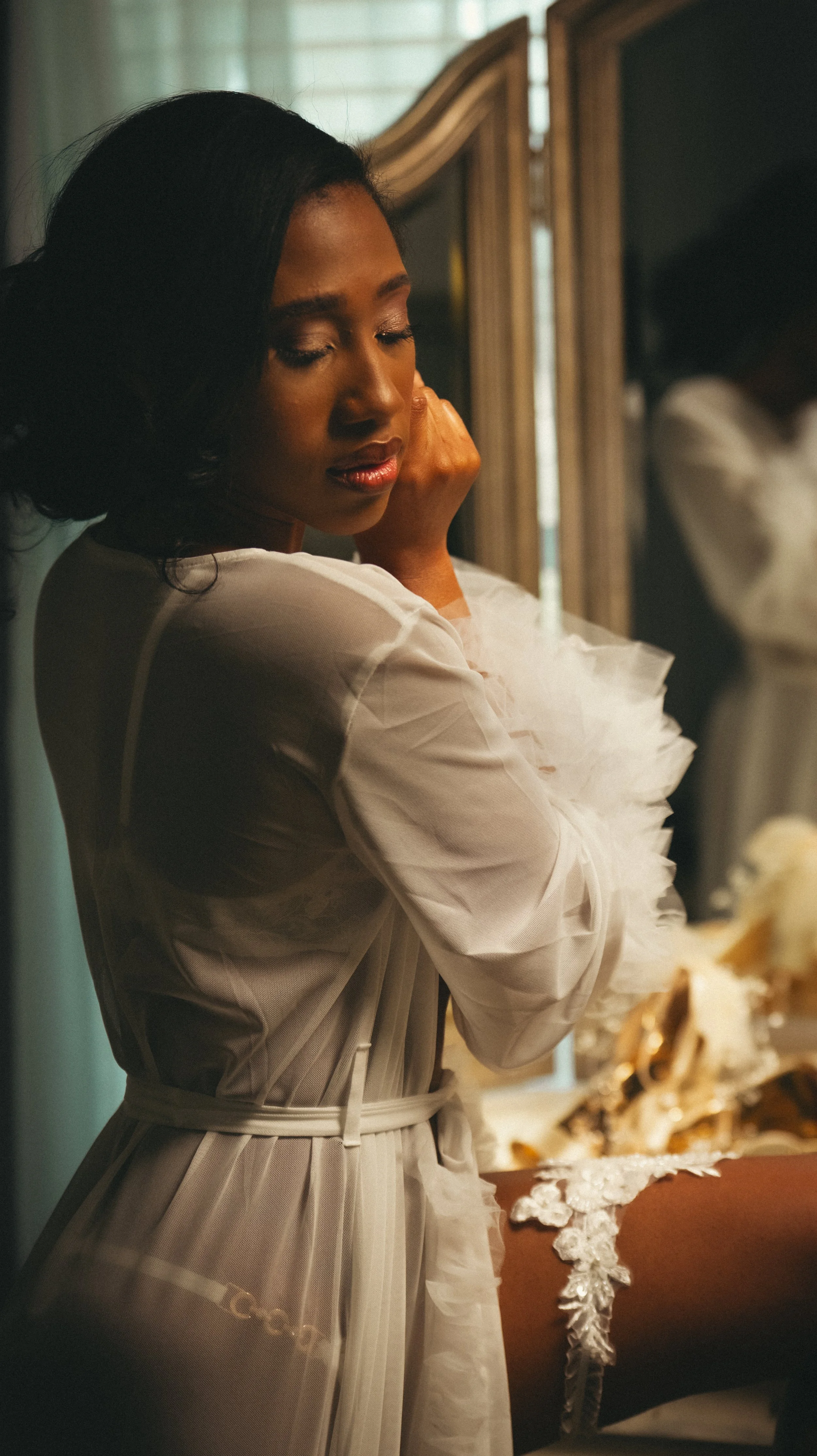 A woman adjusting her hair in front of a mirror, wearing a white sheer robe and lace lingerie, with soft lighting creating a warm, intimate atmosphere.
