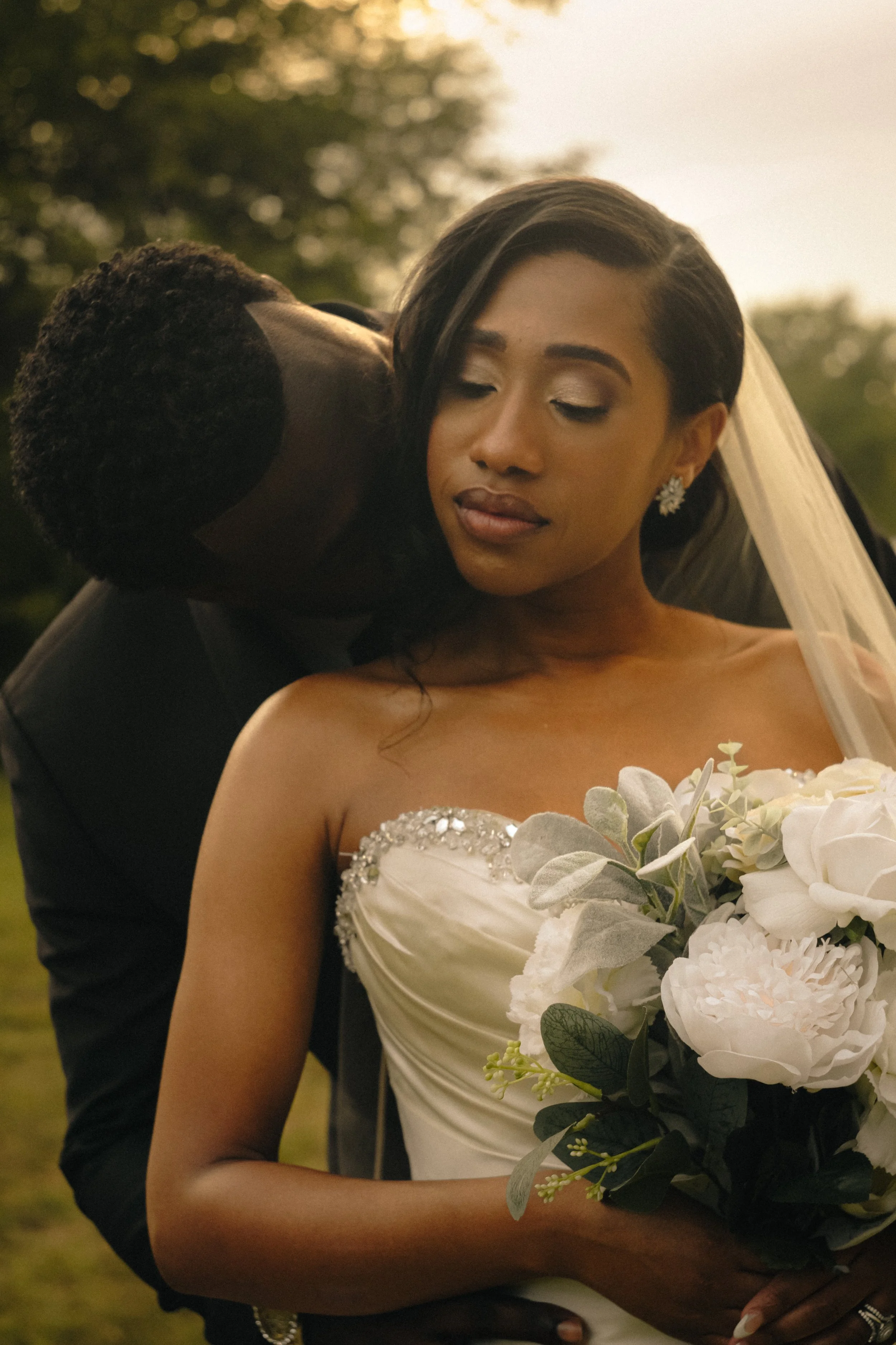 A bride and groom share a tender moment outdoors, with the groom kissing the bride's neck. The bride wears a strapless wedding gown with embellishments and holds a bouquet of white flowers, while the groom is dressed in a black suit.