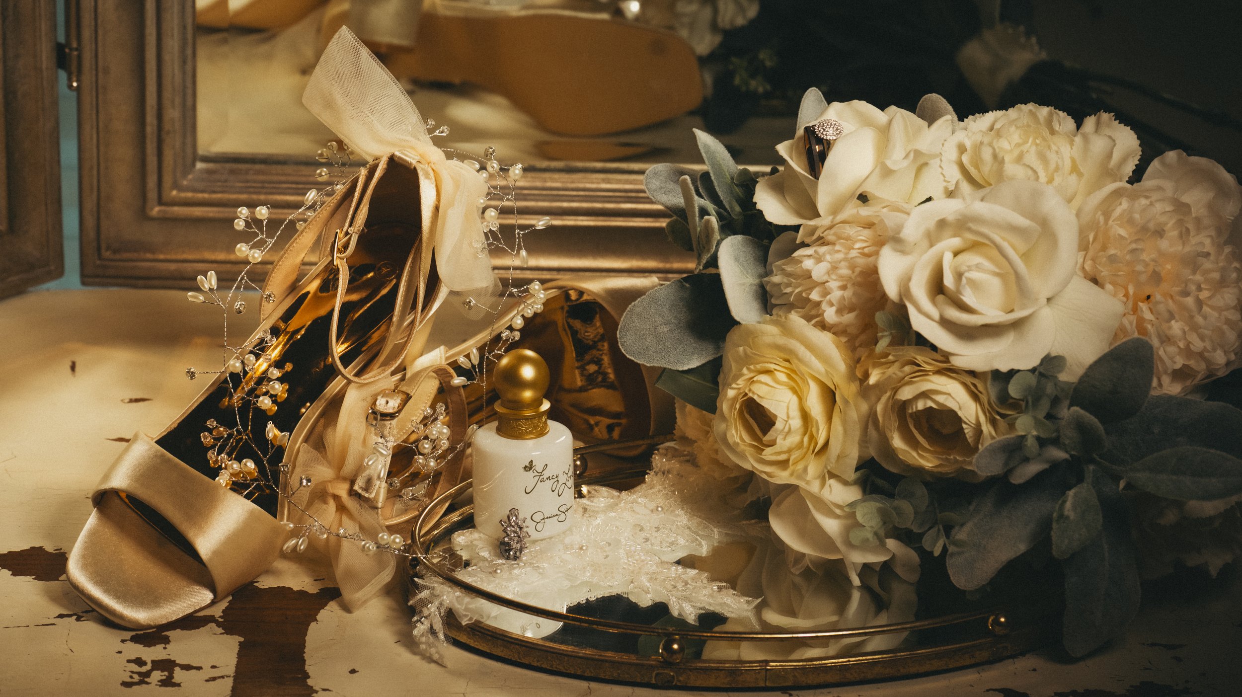 A vintage wooden jewelry box, a satin ribbon, and pearl jewelry on a mirror tray, with a bouquet of cream and white roses and eucalyptus leaves, on a distressed wooden surface.