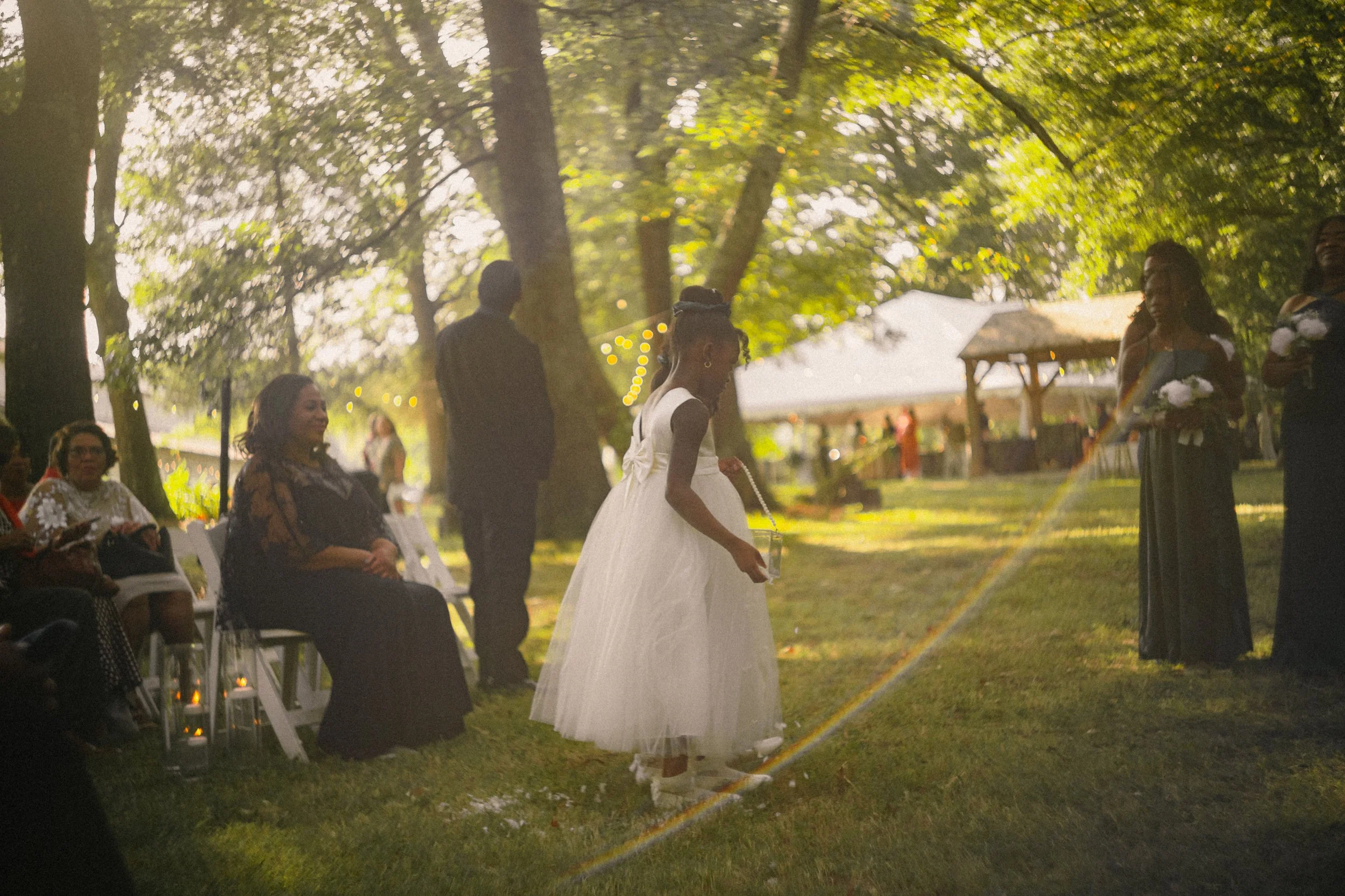A young girl in a white dress holding a drink in an outdoor setting, with adults seated and standing nearby, trees and a pavilion in the background, sunny with warm lighting.
