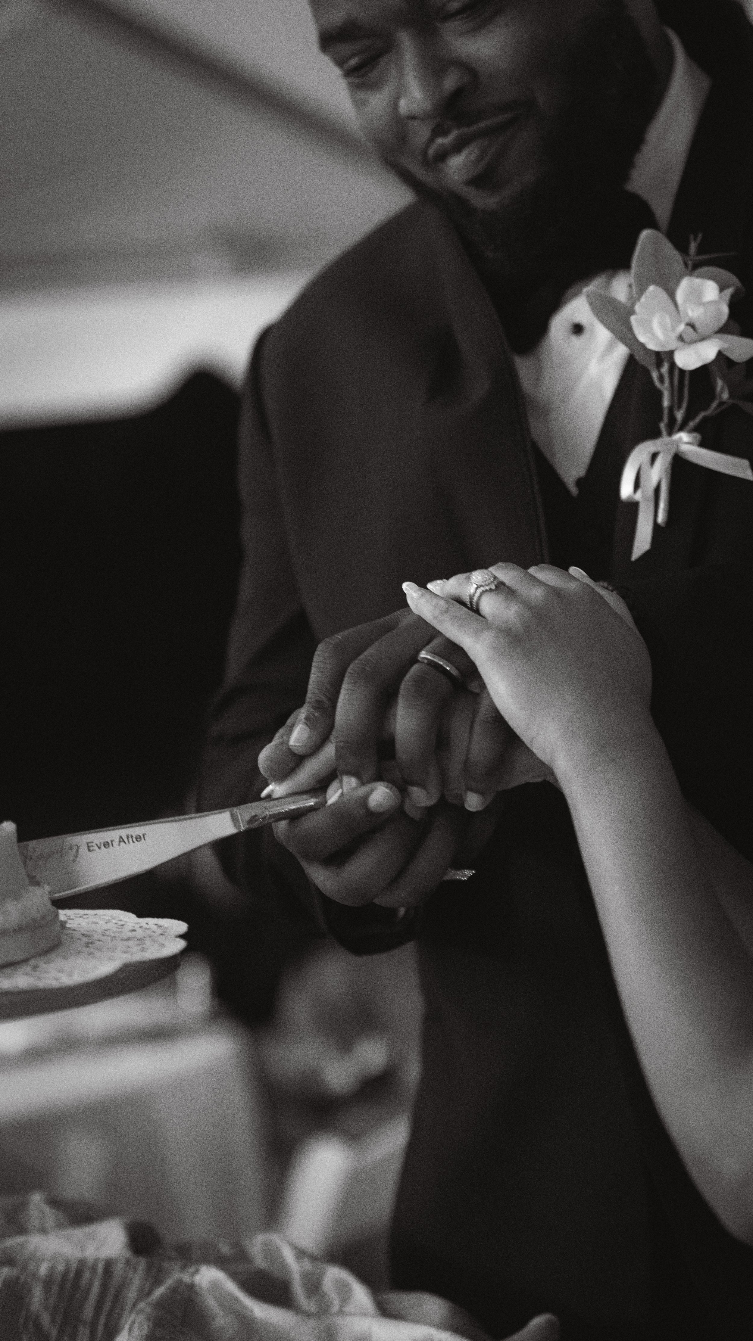 A couple holding hands during a wedding celebration, with the groom wearing a tuxedo and boutonniere, and the bride's hand resting on the groom's hand as they cut a wedding cake together.