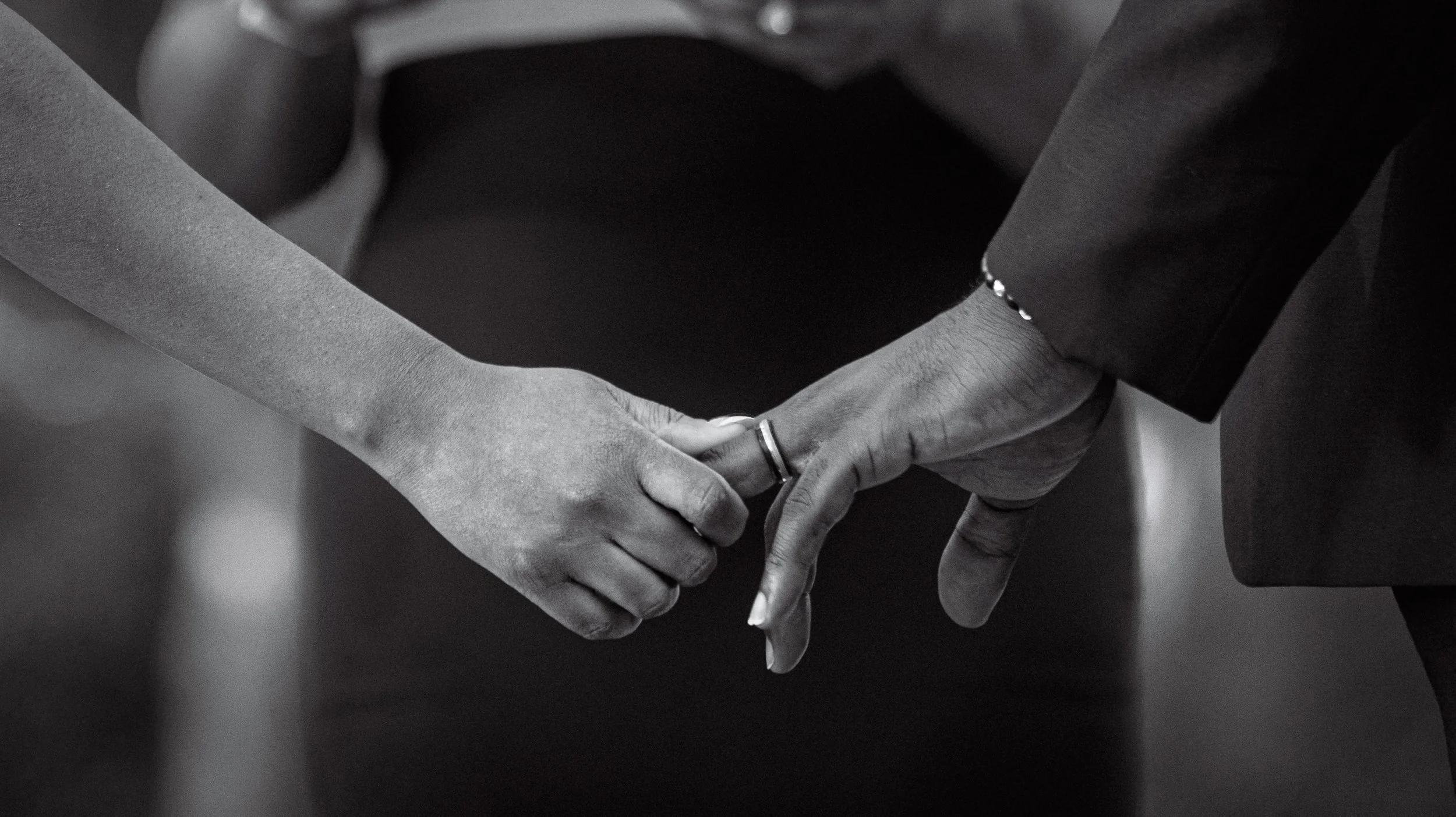 Close-up of two people holding hands, with one person's hand gently grasping the other's fingers, both wearing dark clothing and rings.