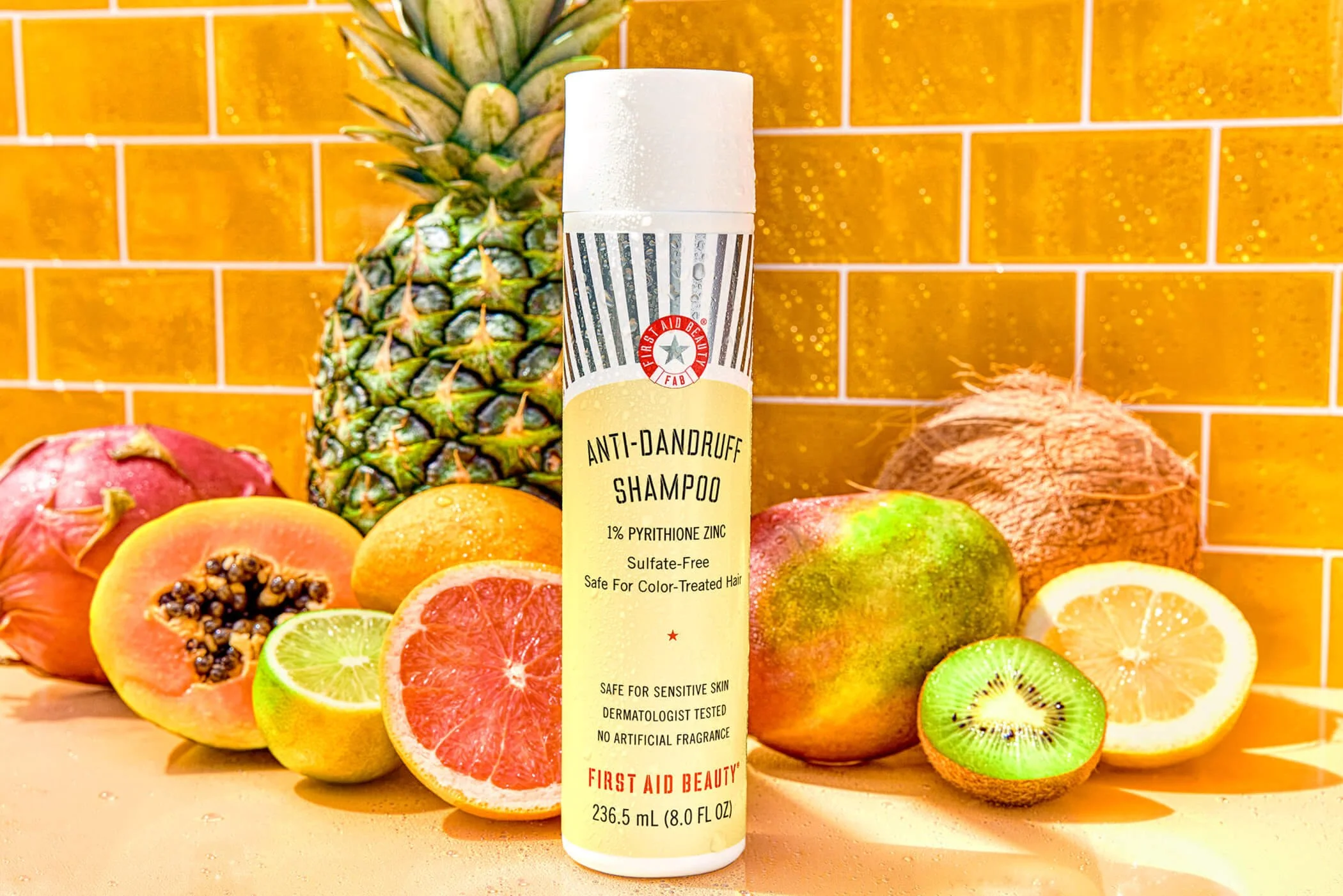 First Aid Beauty anti-dandruff shampoo bottle surrounded by tropical fruits like pineapple, grapefruit, lemon, papaya, coconut, orange, kiwi, and dragon fruit against a yellow tiled background.
