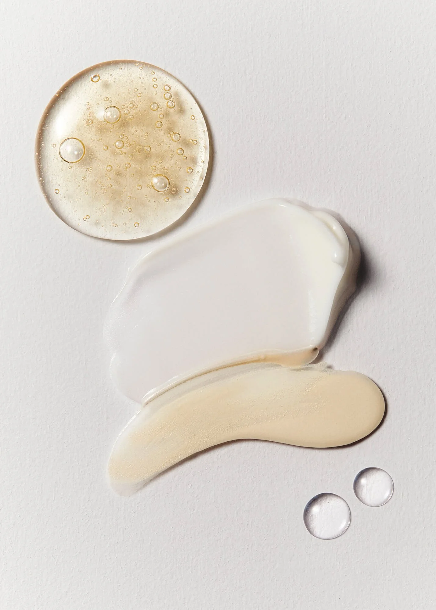 Various skincare product textures on white background: clear gel with bubbles, creamy lotion smear, and small clear droplets.