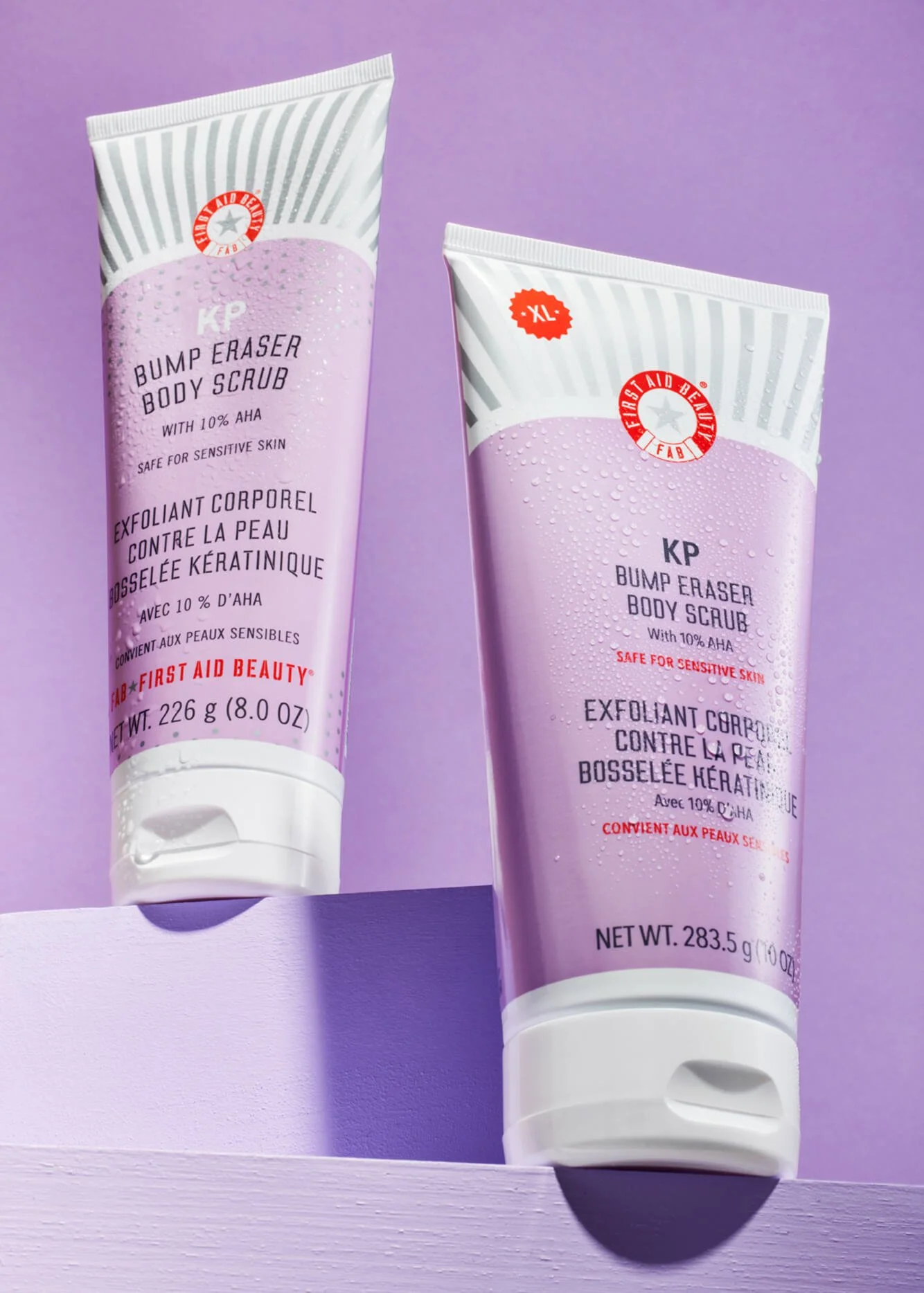 Two tubes of "KP Bump Eraser Body Scrub with 10% AHA" against a purple background, marketed as safe for sensitive skin by First Aid Beauty. One tube is a larger XL size.