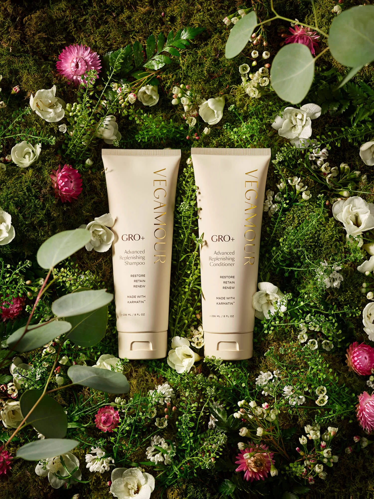 Two bottles of Vegamour GRO+ Advanced Replenishing Shampoo and Conditioner on a moss-covered surface with pink and white flowers.