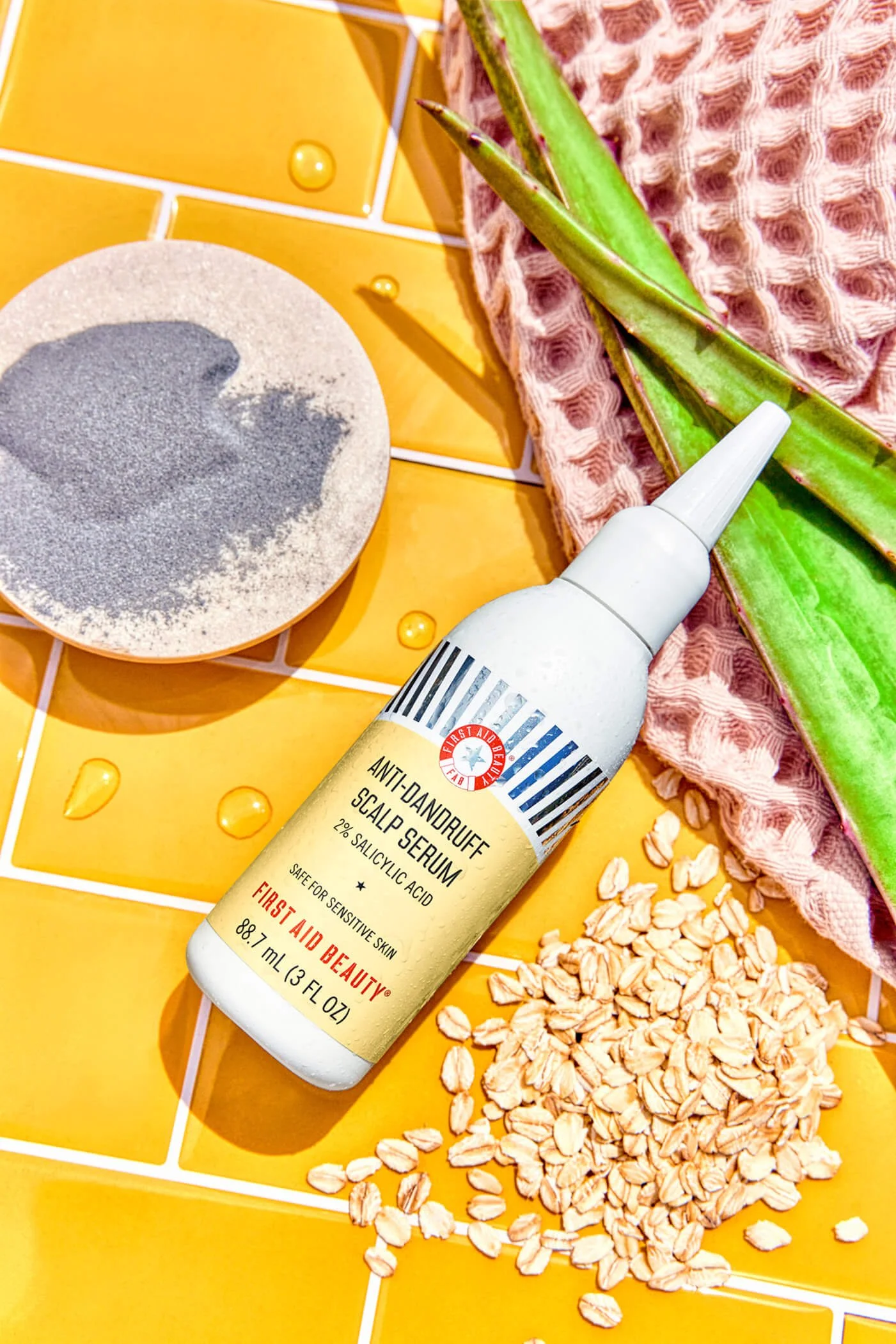 Anti-dandruff scalp serum bottle on a yellow tile background with aloe vera leaves, oats, a pink cloth, and a bowl of powder.