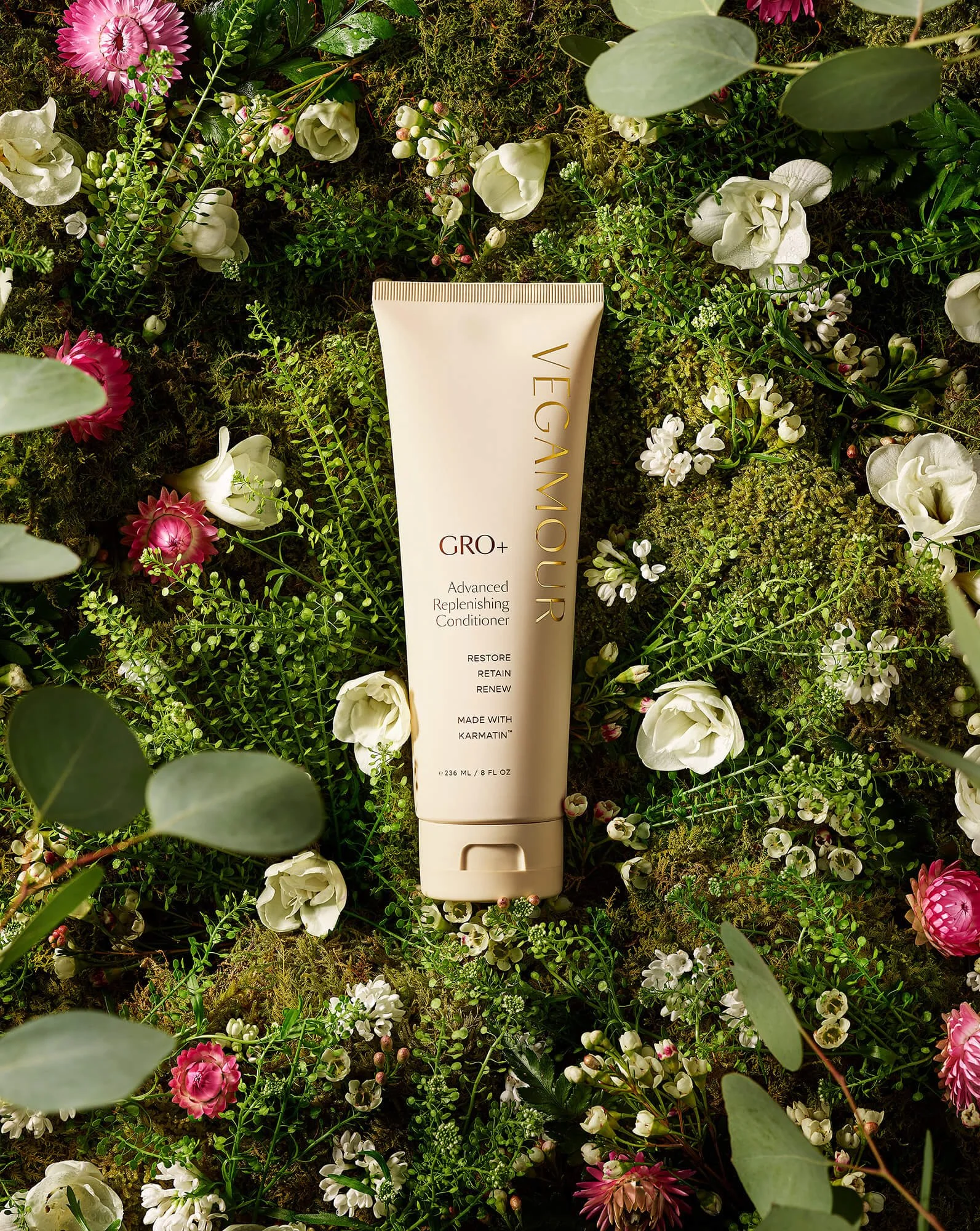 Tube of Vegamour GRO+ Advanced Replenishing Conditioner on a bed of green foliage and white and pink flowers.