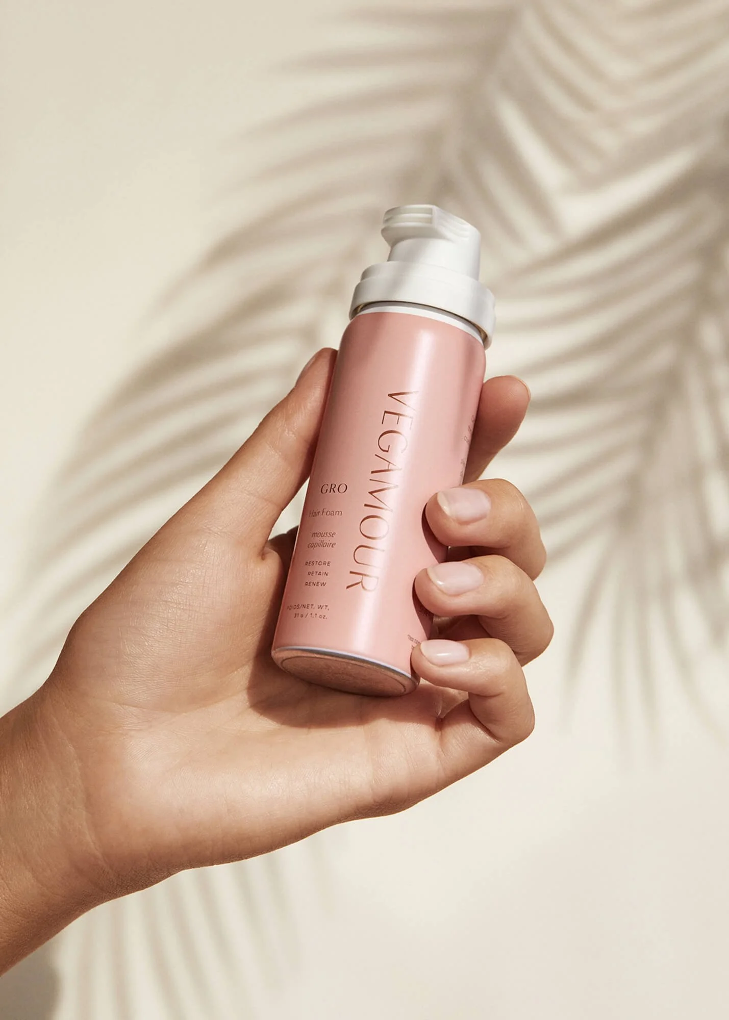 Hand holding a pink Vegamour GRO hair foam bottle with a beige background and shadow of a plant leaf.